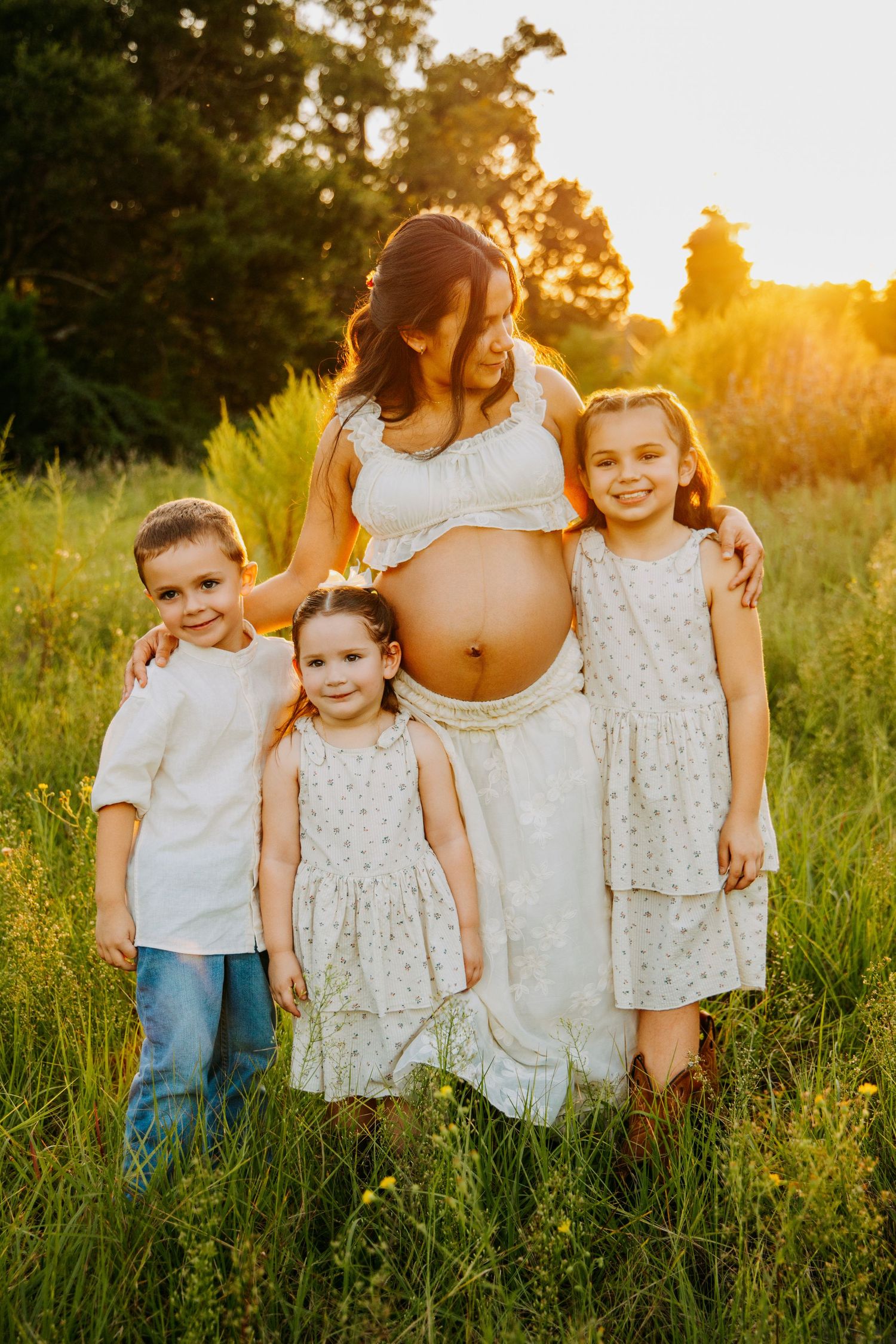 Pregnant woman in white dress poses outdoors at golden hour with three young children in a sunny green meadow.