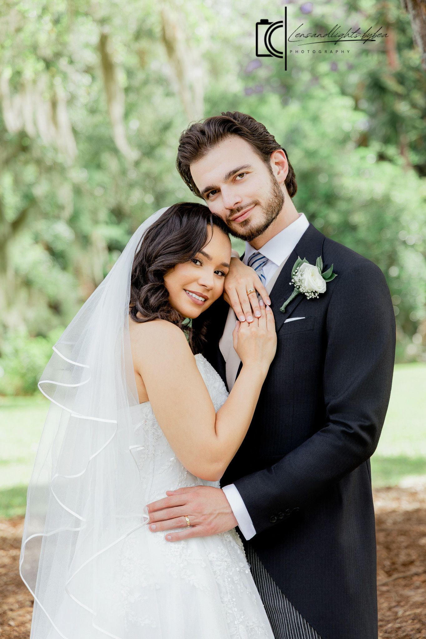 Bride and groom embrace outdoors, she wears veil and lace dress, he wears dark suit with boutonniere.