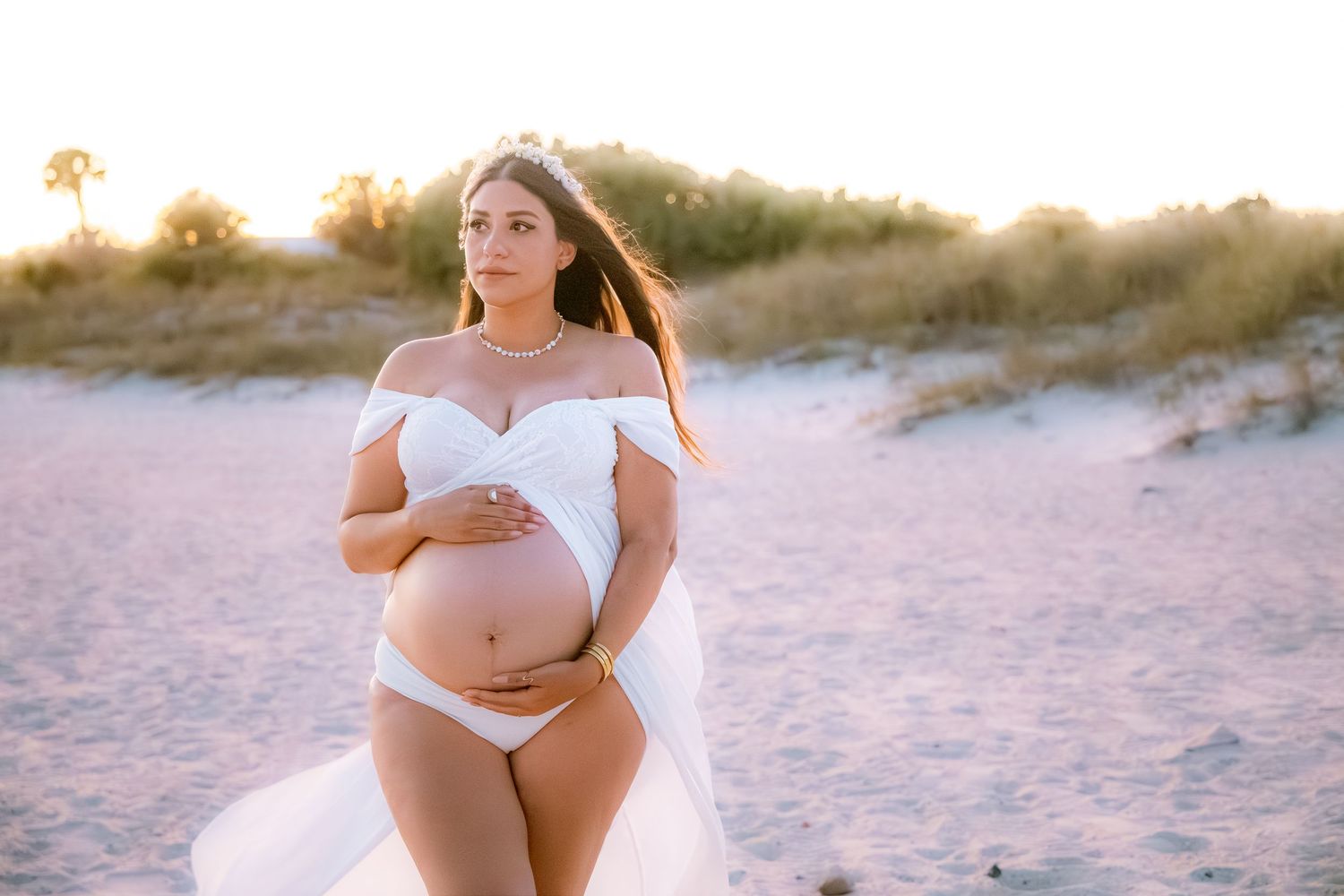 Pregnant woman in white off-shoulder gown cradling baby bump on sandy beach at golden hour sunset.