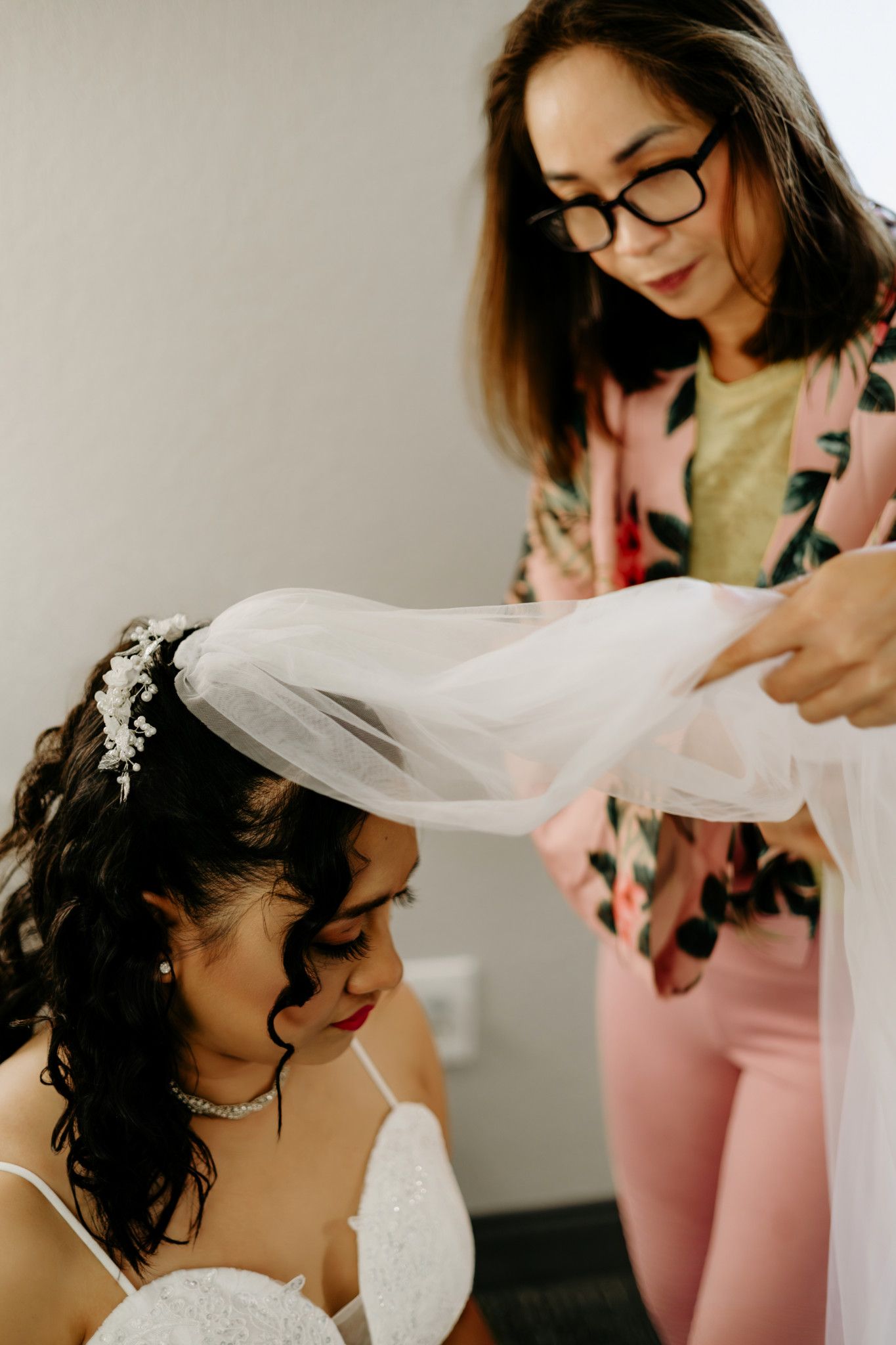 A woman in glasses adjusts a bridal veil on a seated bride wearing a white dress and floral hair accessory.