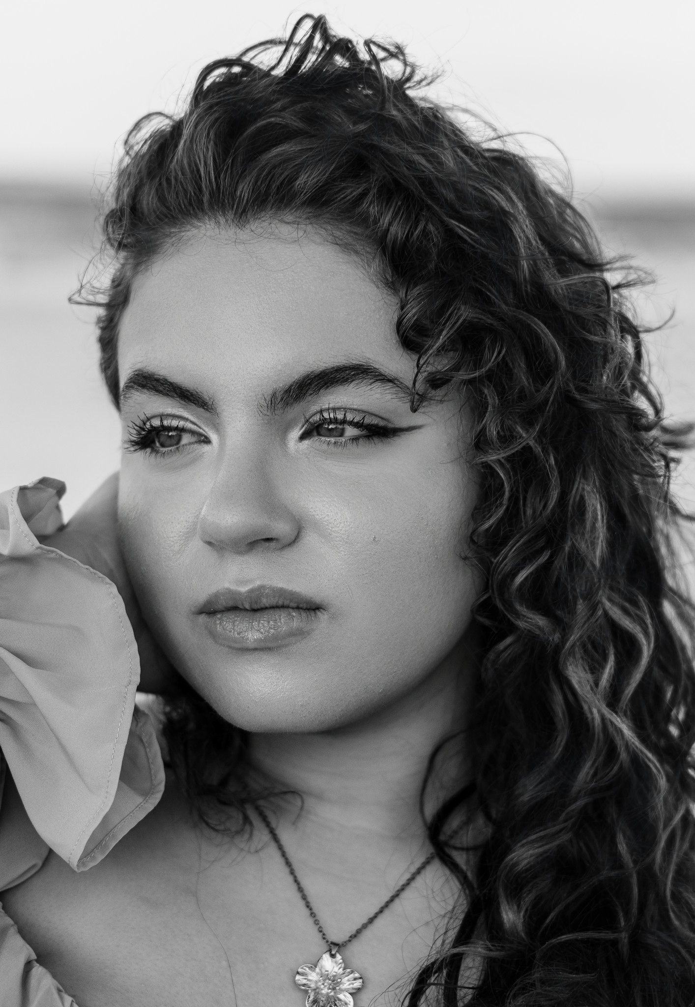 Black and white close-up portrait of a woman with long curly hair, winged eyeliner, and a flower pendant necklace gazing off to the side.