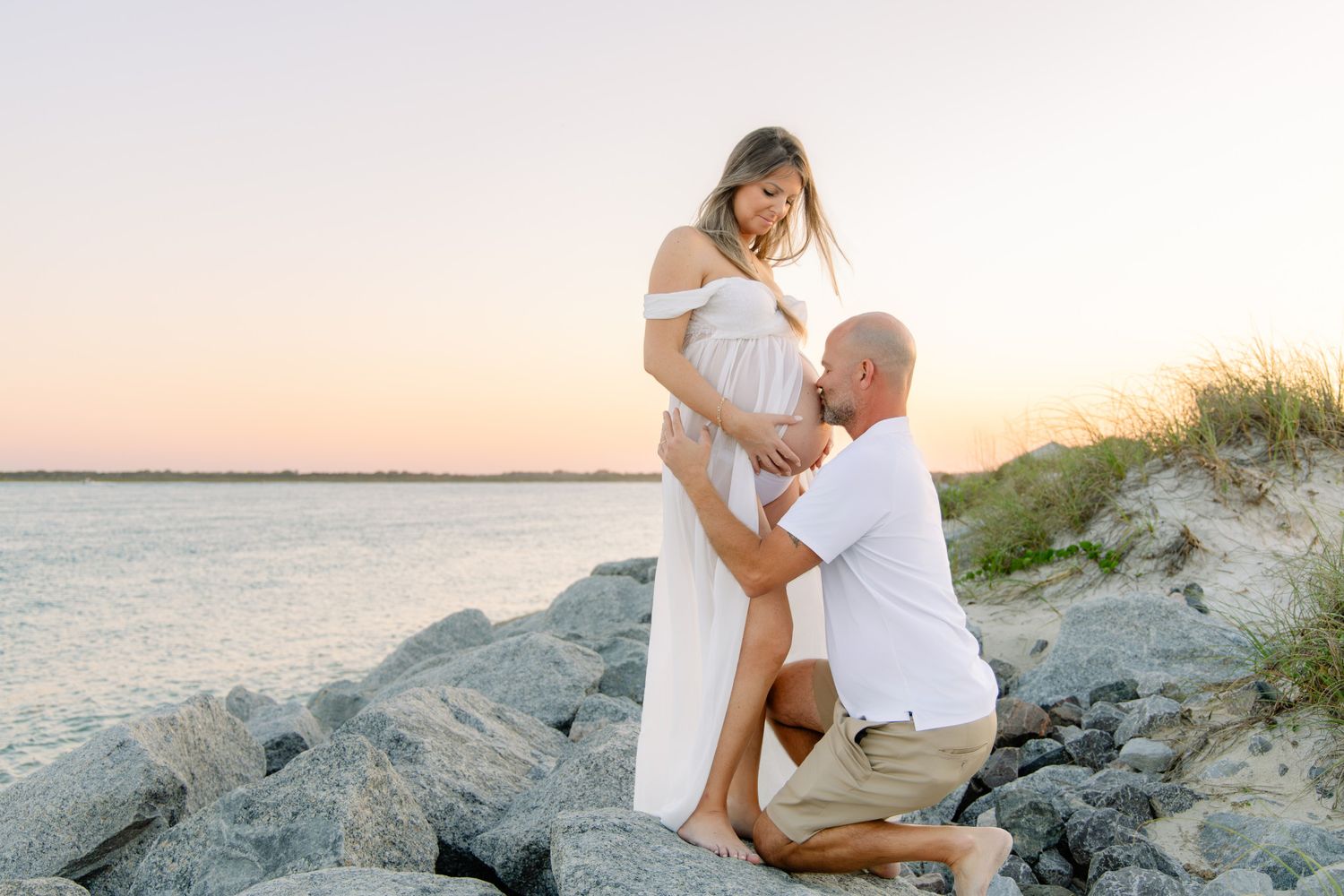 Couple at beach sunset, man kneeling touching pregnant woman's belly on rocky shore.