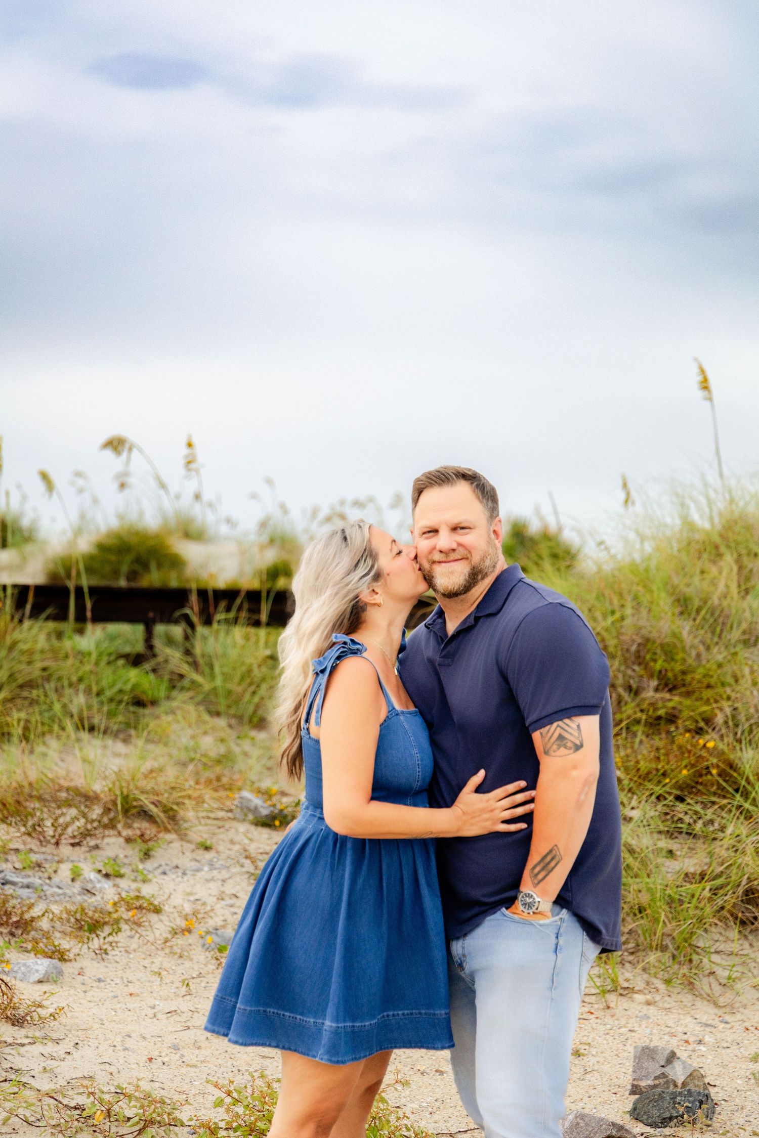 A couple embraces and kisses on a sandy beach, woman in blue dress, man in navy polo with tattoos, sea oats in background.