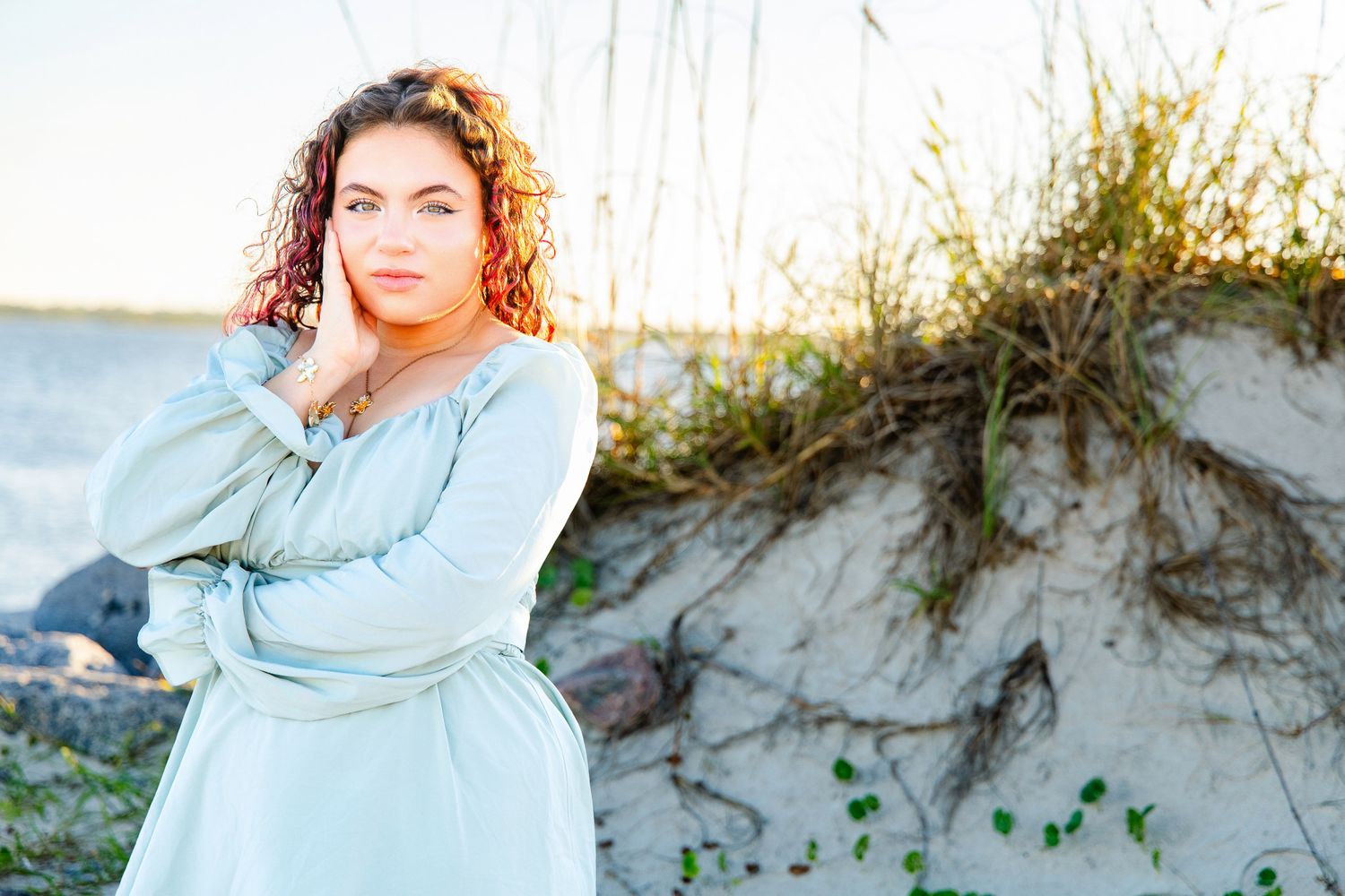 Young woman with curly auburn hair in a flowing sage green dress poses with her hand on her cheek beside a sandy dune with sea oats.