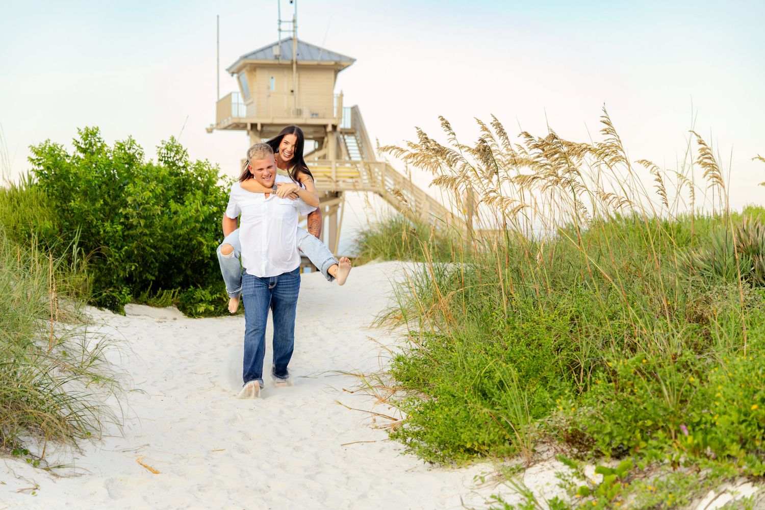 Man giving woman piggyback ride on sandy beach path with sea oats and lifeguard tower behind them.