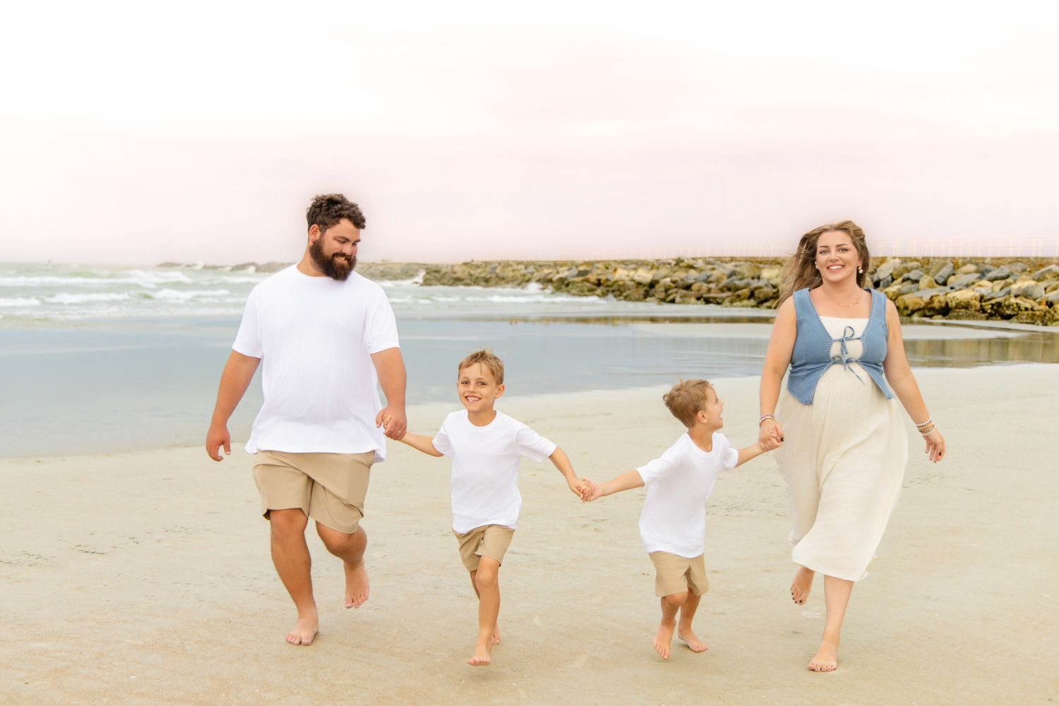 Happy family of four holding hands and running on the beach at sunset near rocky shoreline.