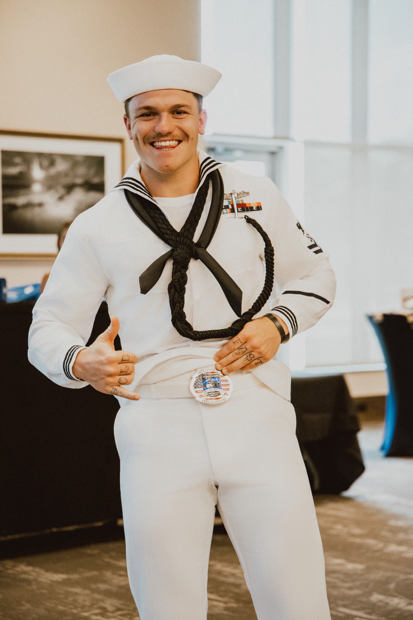 Smiling Navy sailor in white dress uniform with black neckerchief and white dixie cup hat posing indoors.