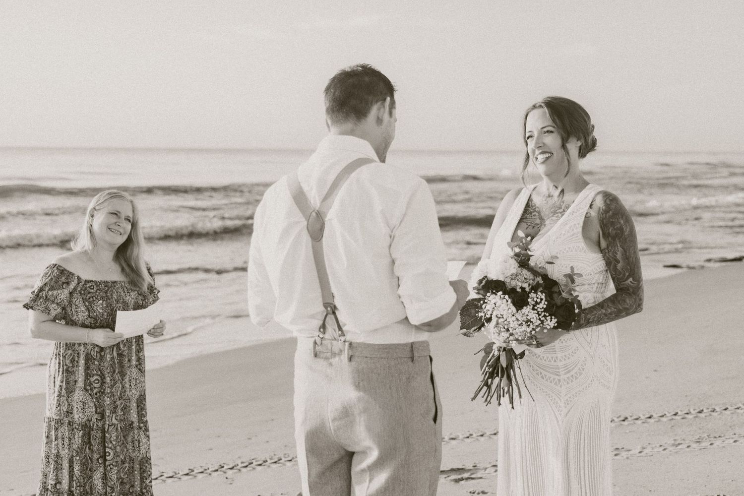 Black and white beach wedding ceremony with bride holding bouquet, groom in suspenders, and officiant by the ocean.