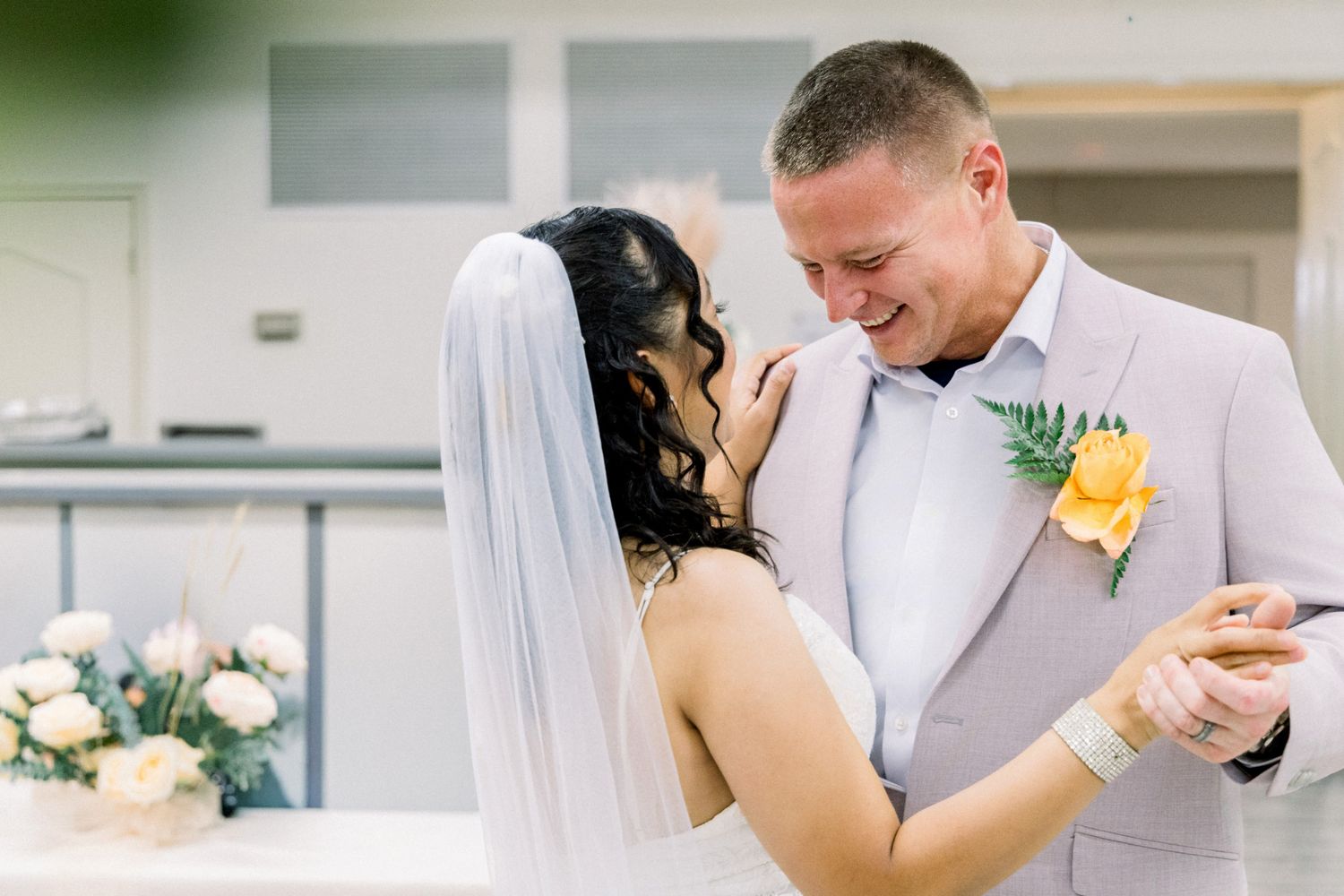 Bride and groom share first dance, smiling intimately, bride in white veil, groom in light suit with yellow boutonniere.
