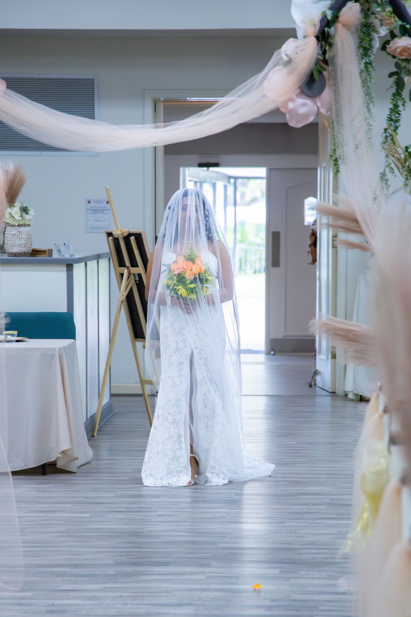 Bride in white lace gown and veil holding bouquet walks toward entrance of decorated wedding venue.