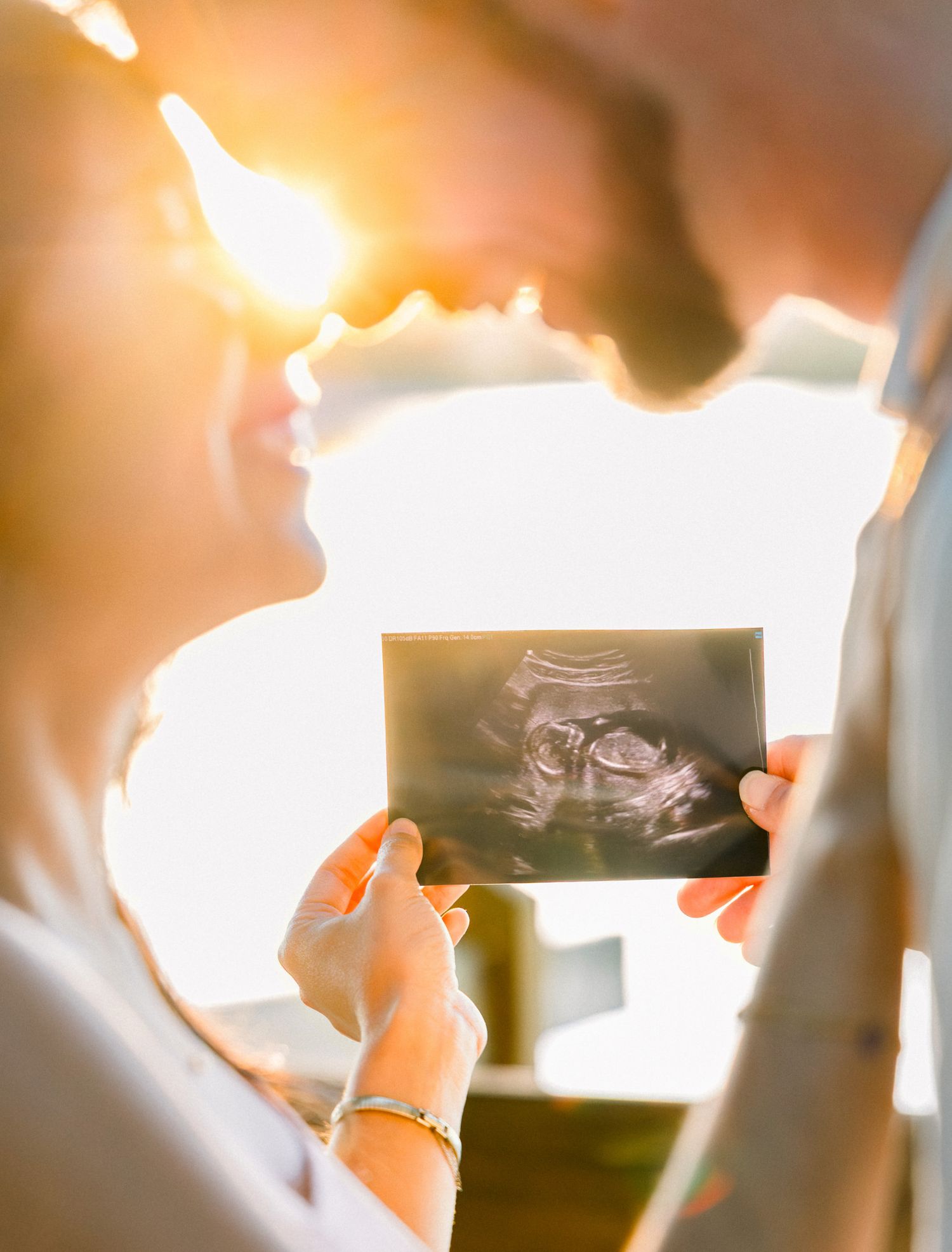 Couple holding ultrasound photo in golden sunlight, sharing a tender moment as they anticipate their new baby.
