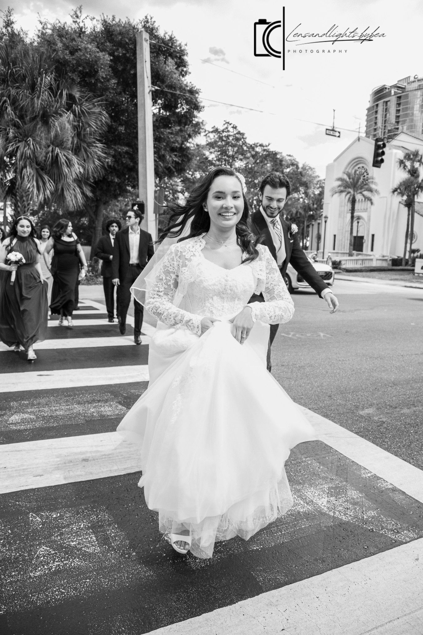Bride in lace wedding gown crossing street with wedding party in black and white photo.