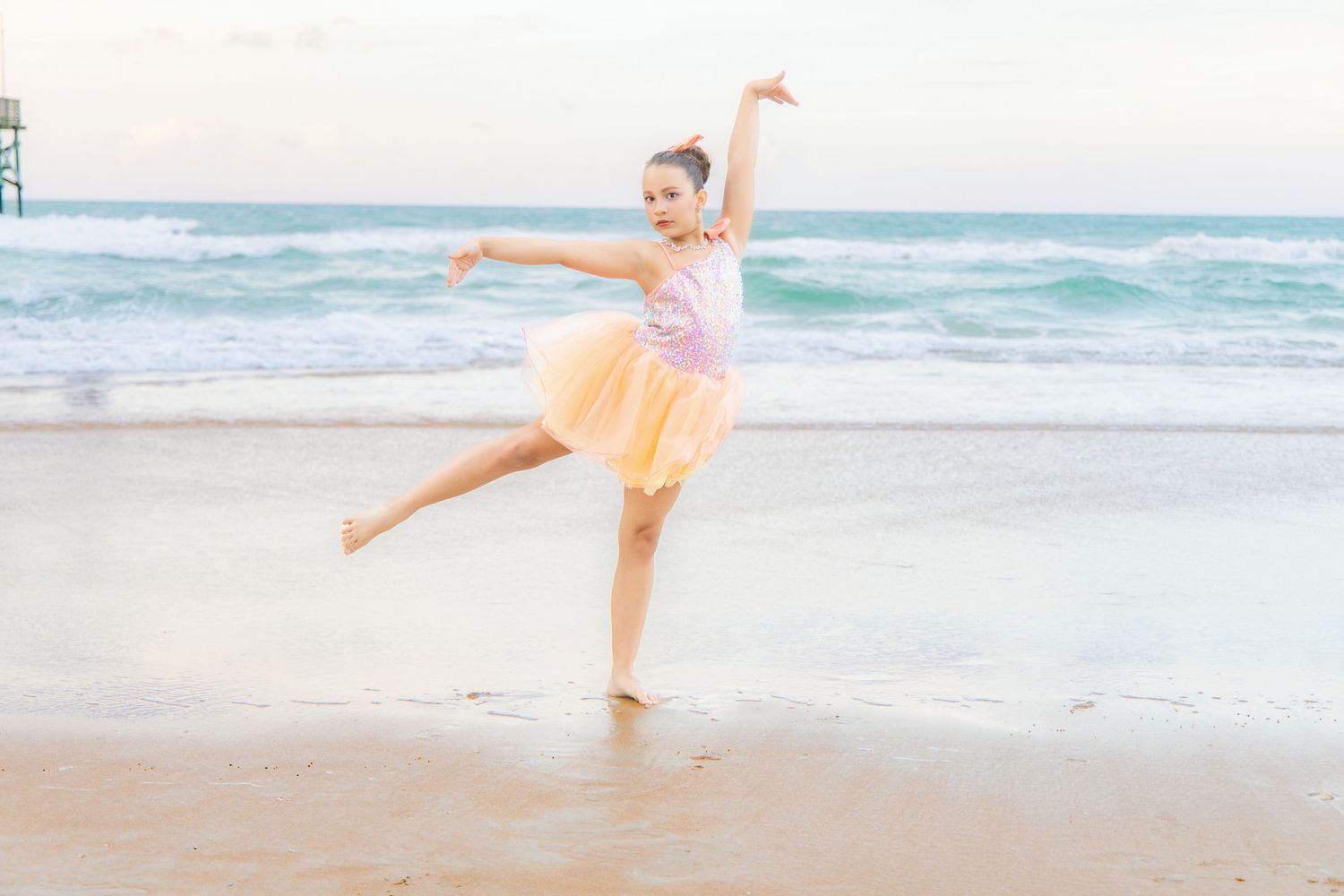 Young ballet dancer performs graceful arabesque pose on sandy beach with turquoise ocean waves in background.