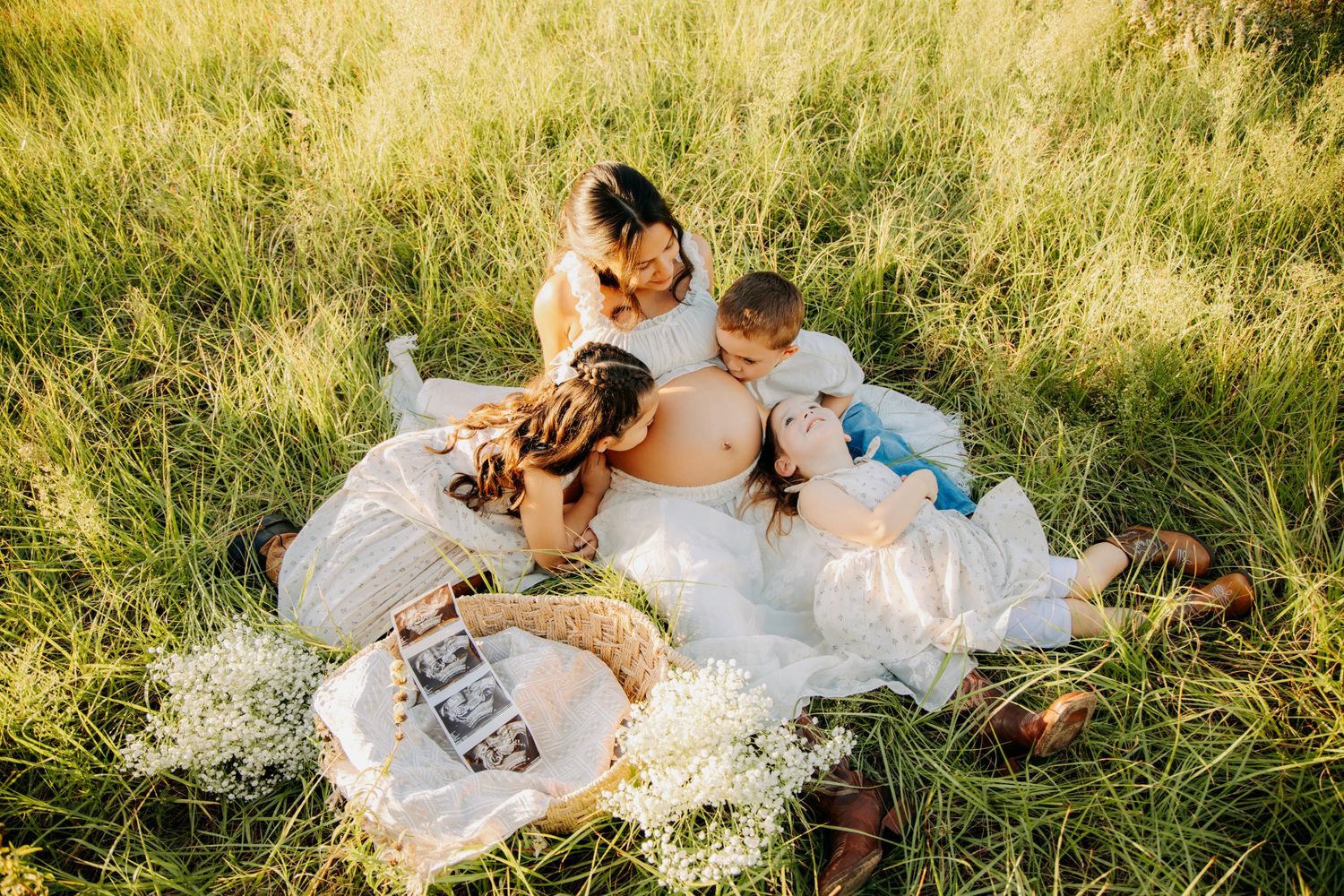 Pregnant woman relaxing in green field with family, ultrasound photos displayed on blanket during maternity photoshoot.