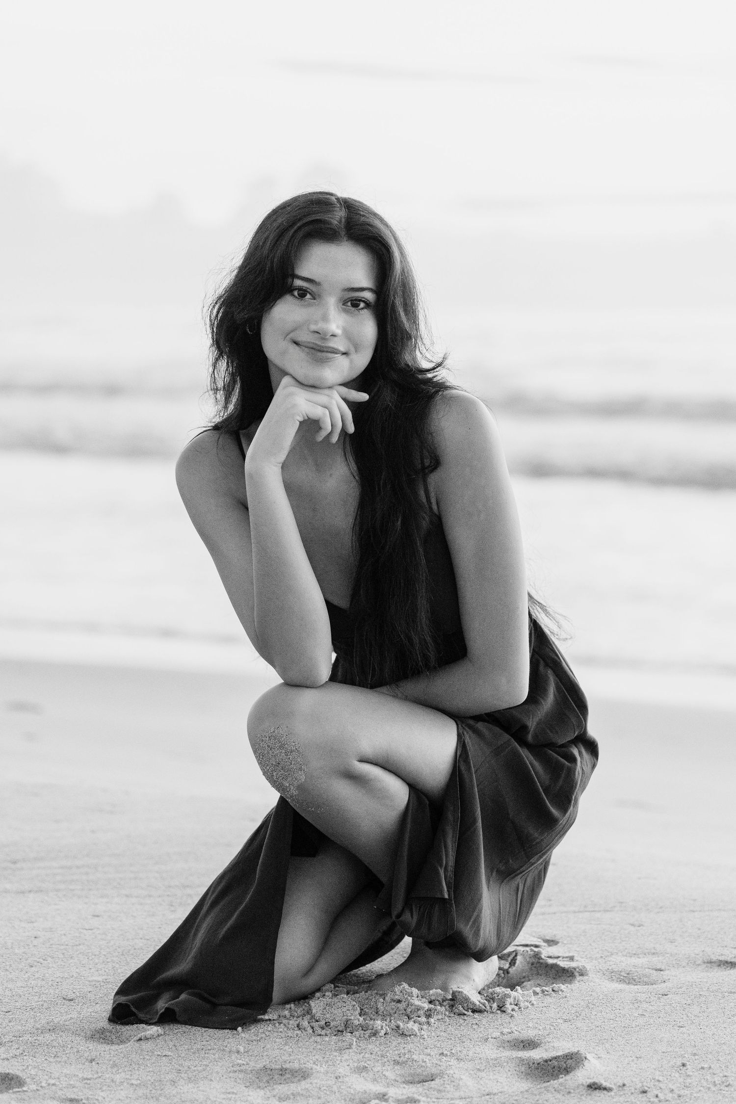 Black and white photo of a young woman in a dark dress crouching on a sandy beach with ocean waves in the background.