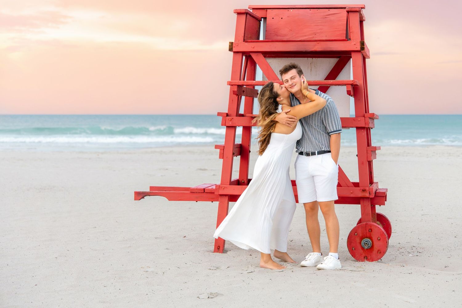 Couple embracing by a red lifeguard stand on a sandy beach at sunset with ocean waves in the background.