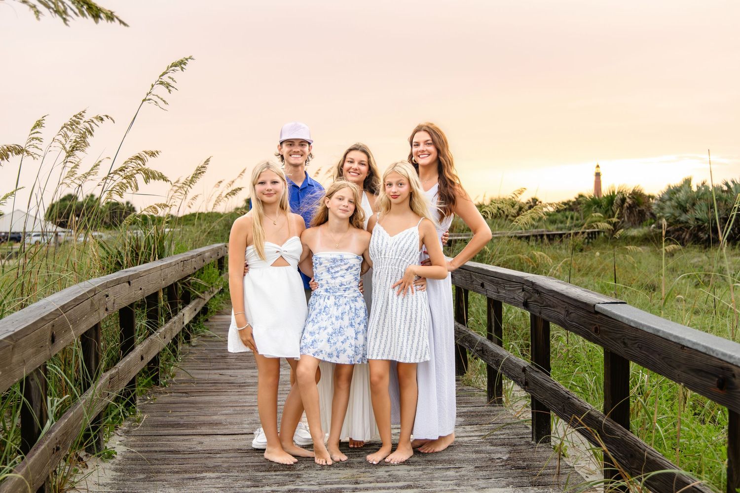 Family of six posing on a wooden boardwalk at sunset near coastal dunes and a lighthouse.