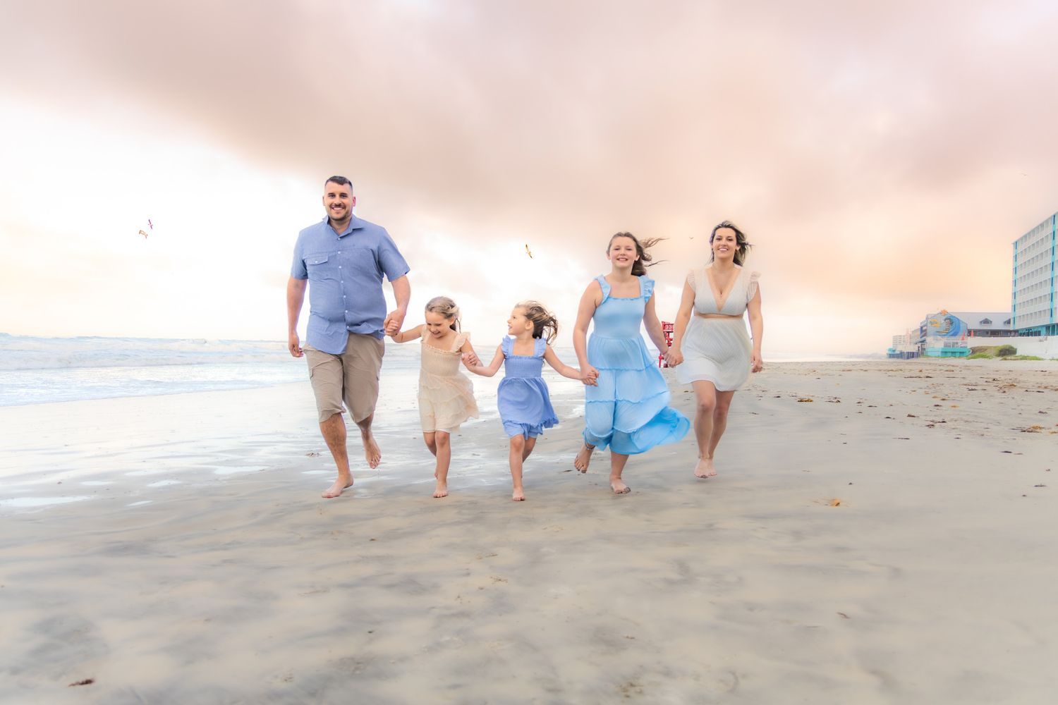 Happy family of five running on a sandy beach at sunset, wearing coordinating blue and white outfits.