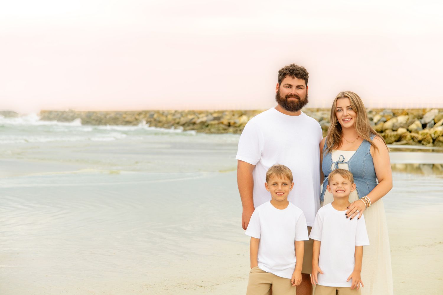 A family of four poses together on a beach near rocky jetty, parents with two young boys in matching outfits.