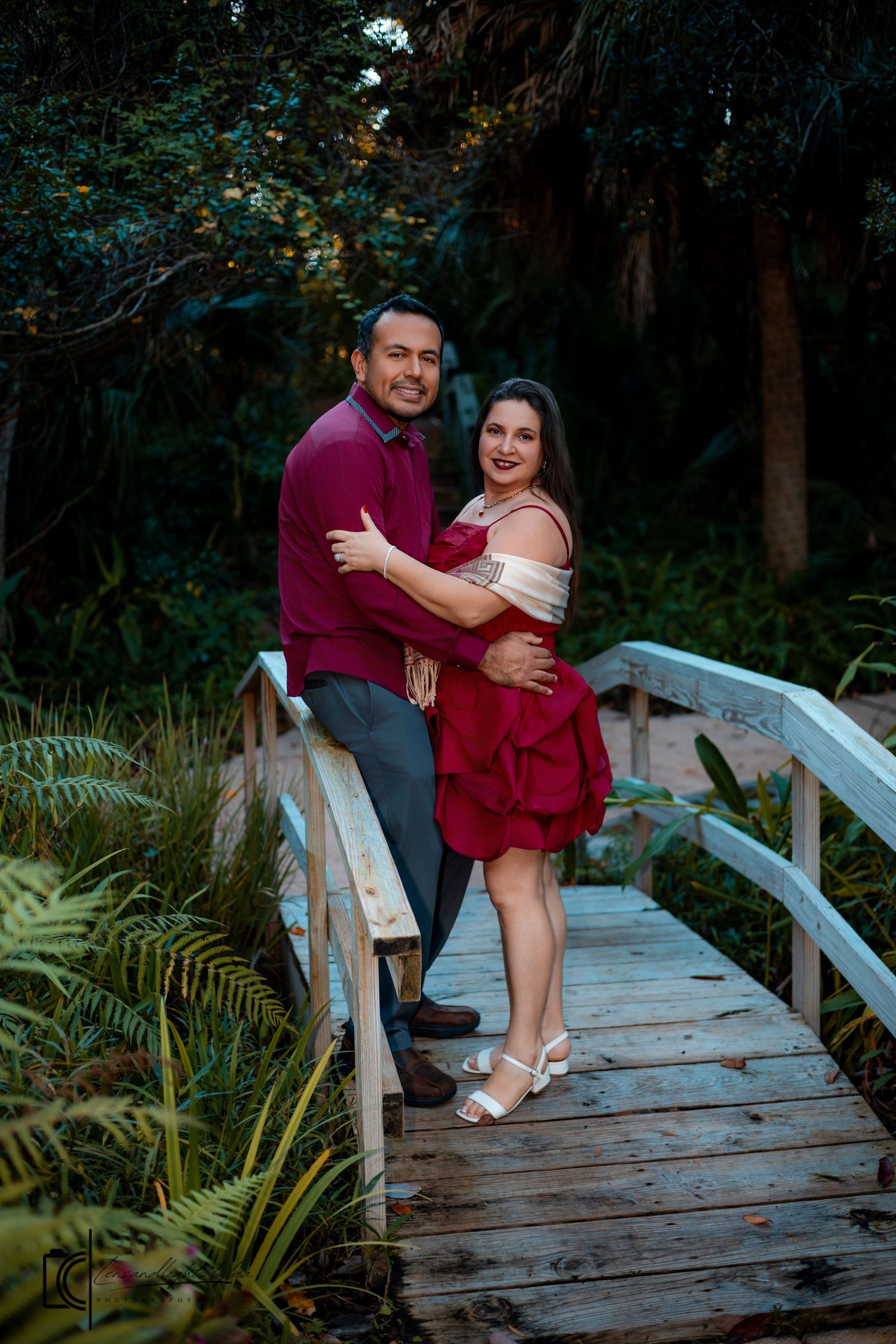 A couple sits together on a wooden boardwalk surrounded by lush ferns and greenery in a romantic outdoor setting.