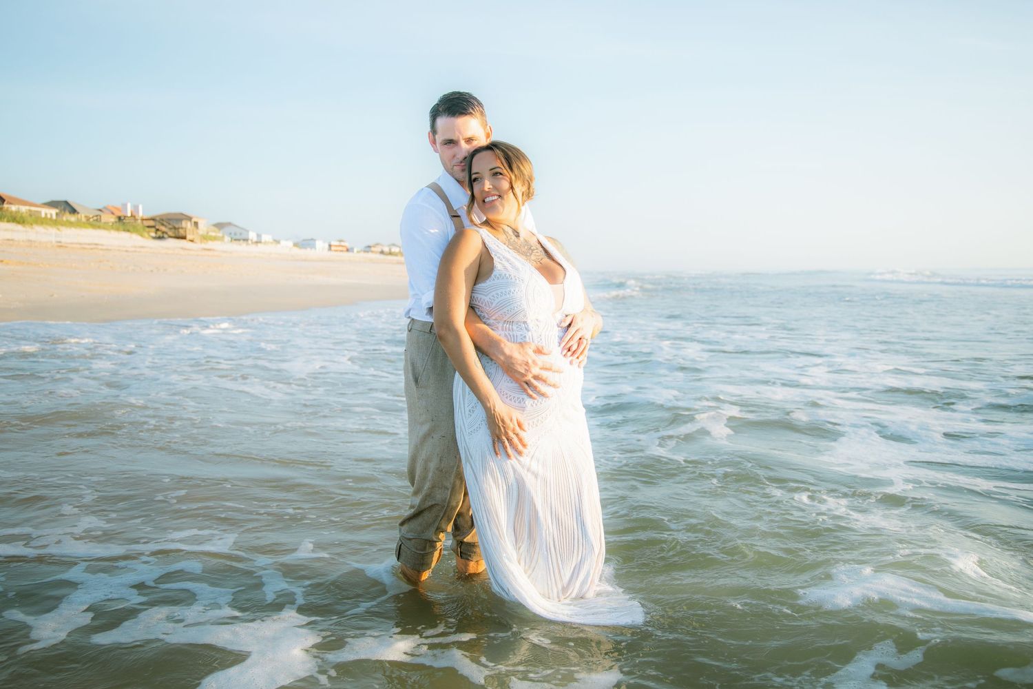 Pregnant couple embracing in ocean waves during a beach maternity photo session at golden hour.
