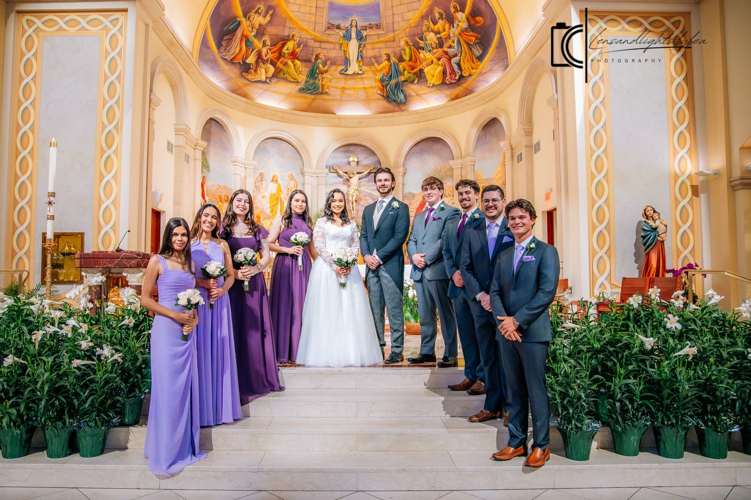 Wedding party poses in church aisle, bridesmaids in purple gowns, groomsmen in gray suits, bride and groom centered.