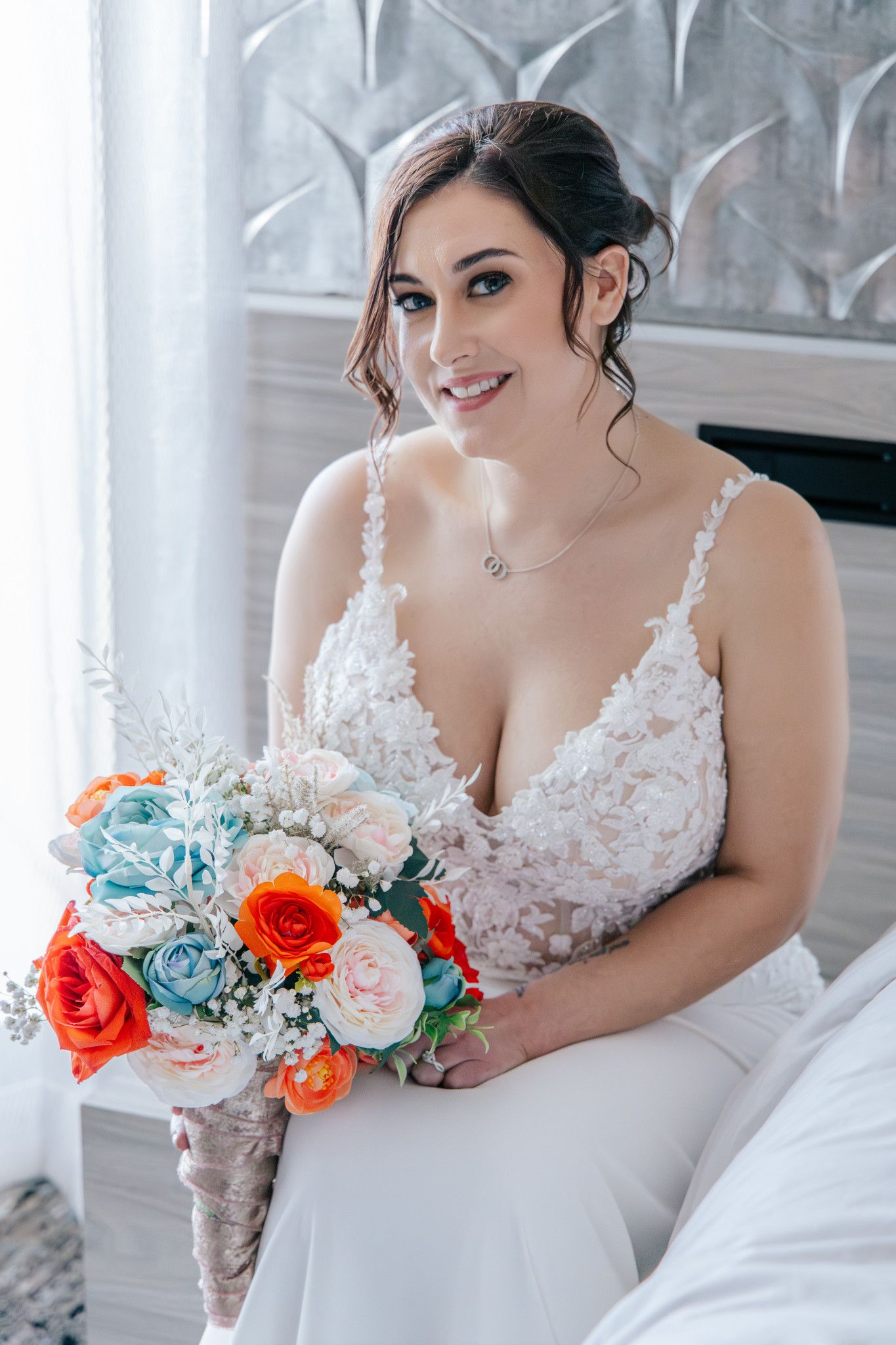Smiling bride in a white lace gown holding a colorful bouquet of orange, pink, and blue roses indoors.