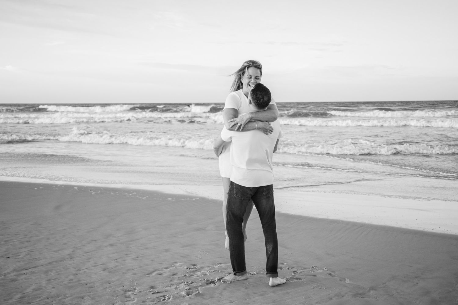 Black and white photo of a couple embracing on a sandy beach with ocean waves in the background.