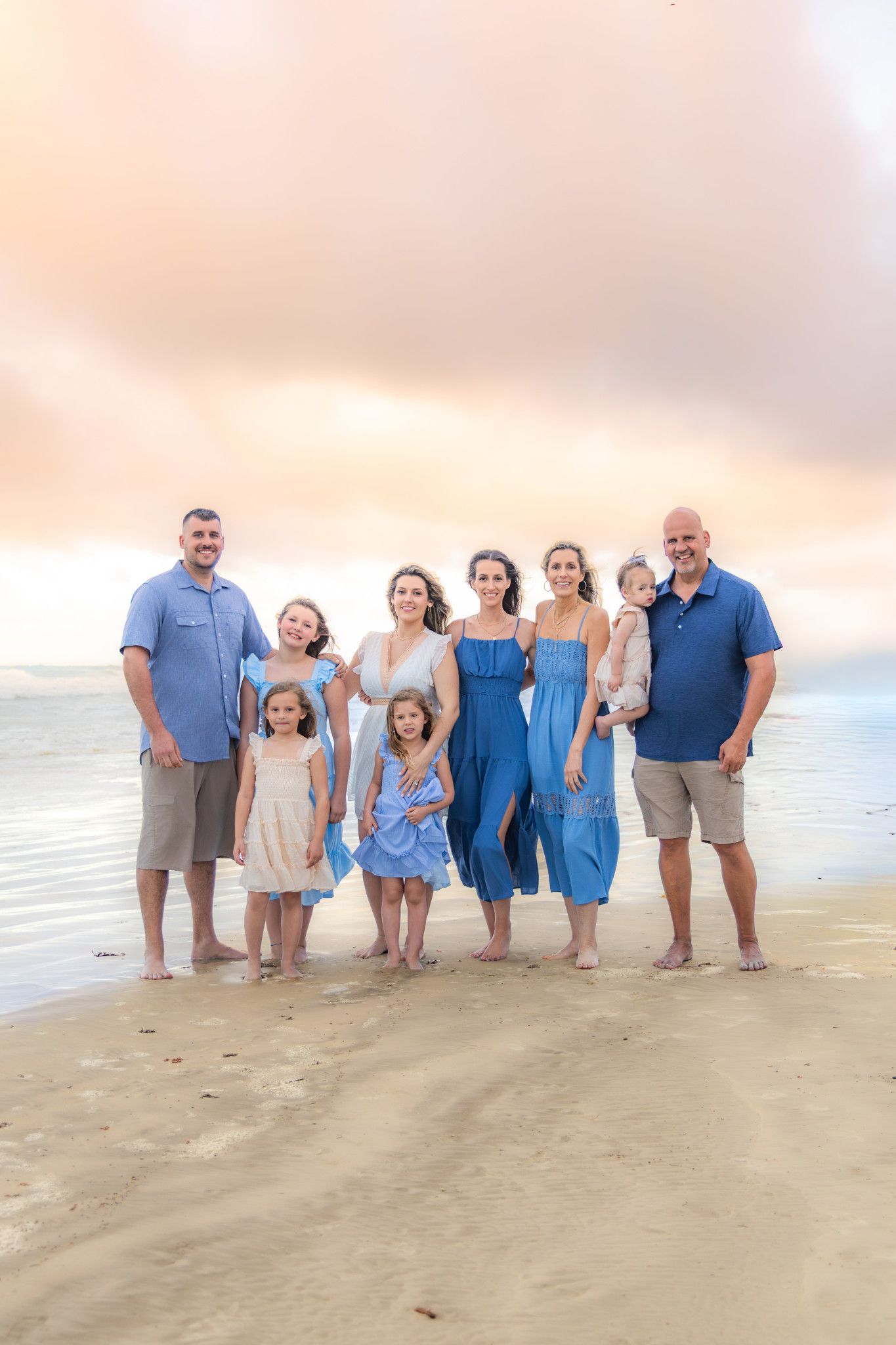 Family of eight poses on a sandy beach at sunset, wearing coordinating blue and neutral outfits, with calm ocean waves behind them.