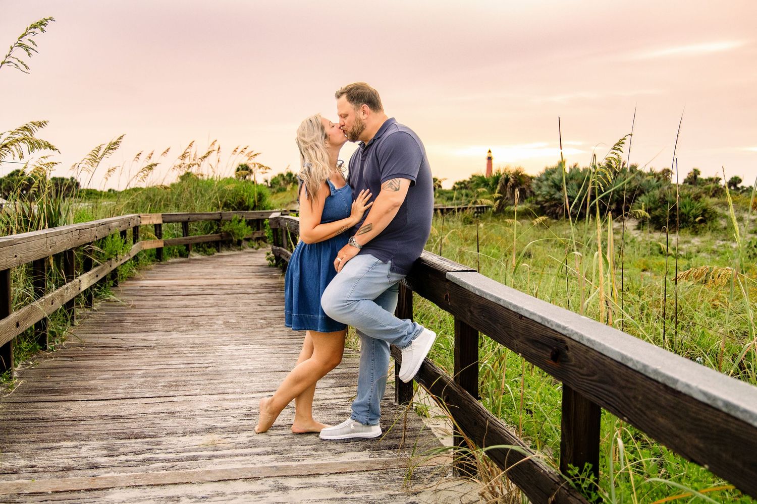 Couple sharing a romantic kiss on a wooden boardwalk surrounded by lush greenery at sunset.
