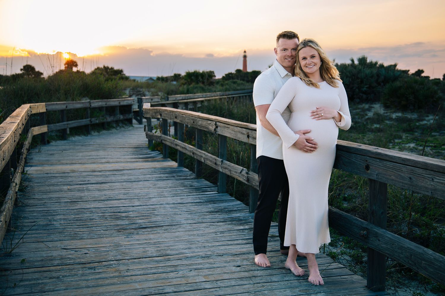 A couple poses together on a wooden boardwalk at sunset during a maternity photo session.