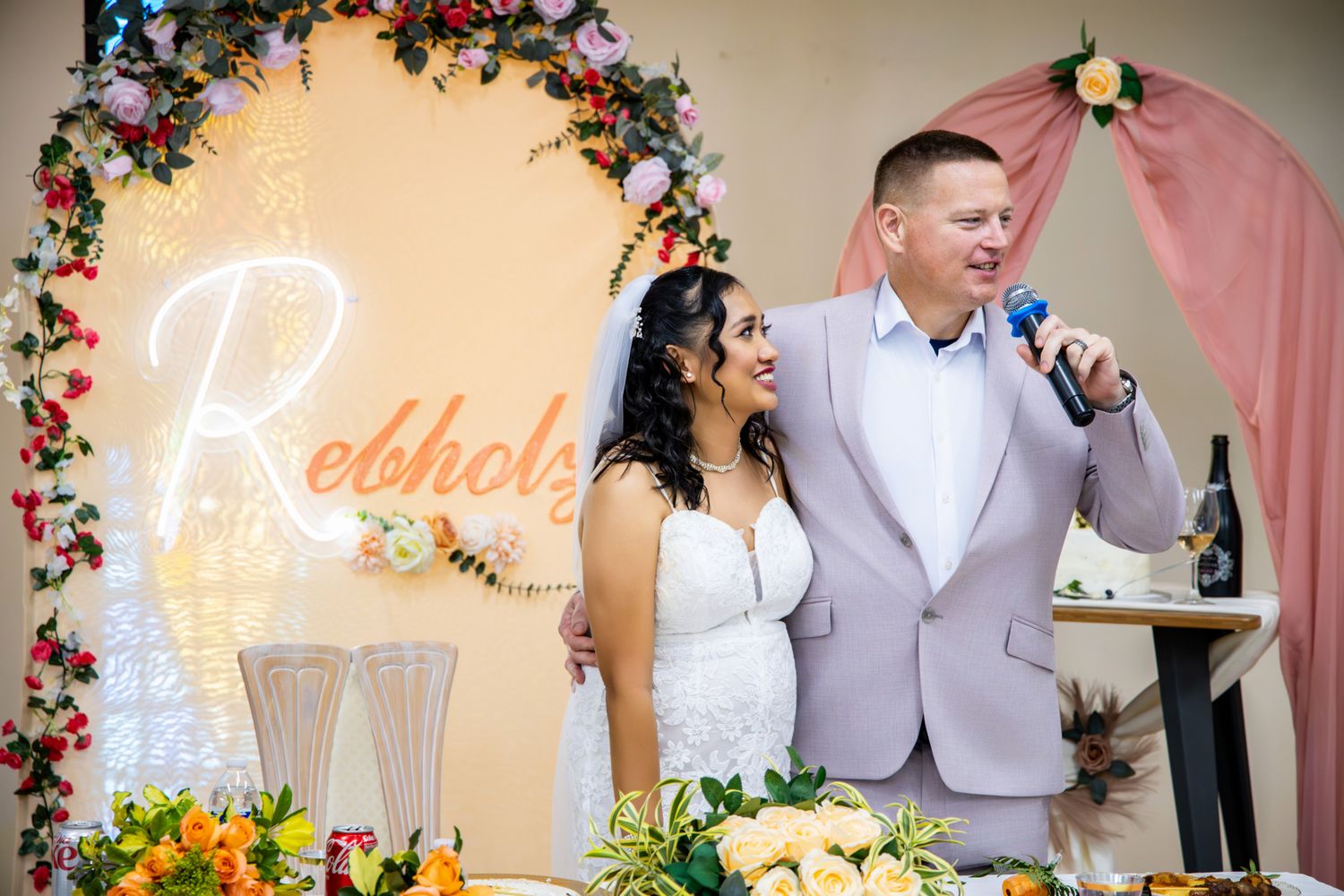 Bride and groom smiling at wedding reception, standing before floral arch with glowing Rebholz sign.