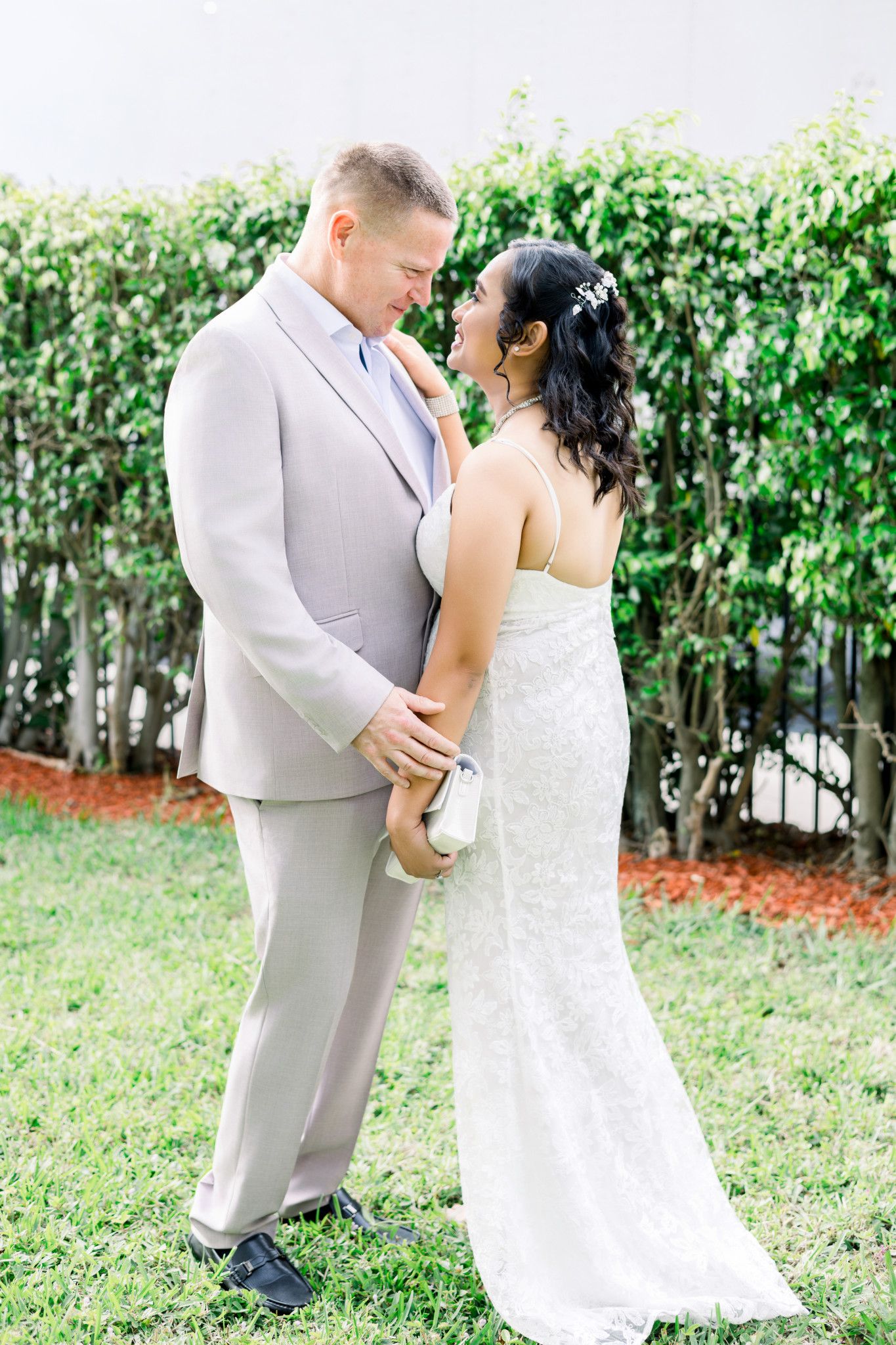 A bride and groom share a romantic moment outdoors, with the groom in a gray suit and bride in a white lace gown.