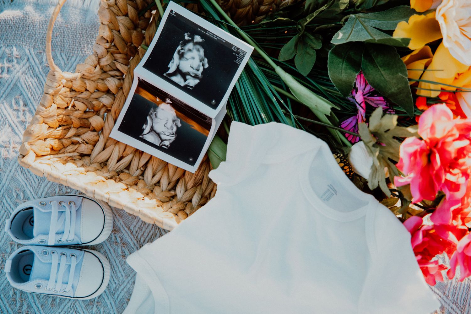 Flat lay of baby ultrasound photos, tiny sneakers, white onesie, and colorful flowers in a wicker basket.