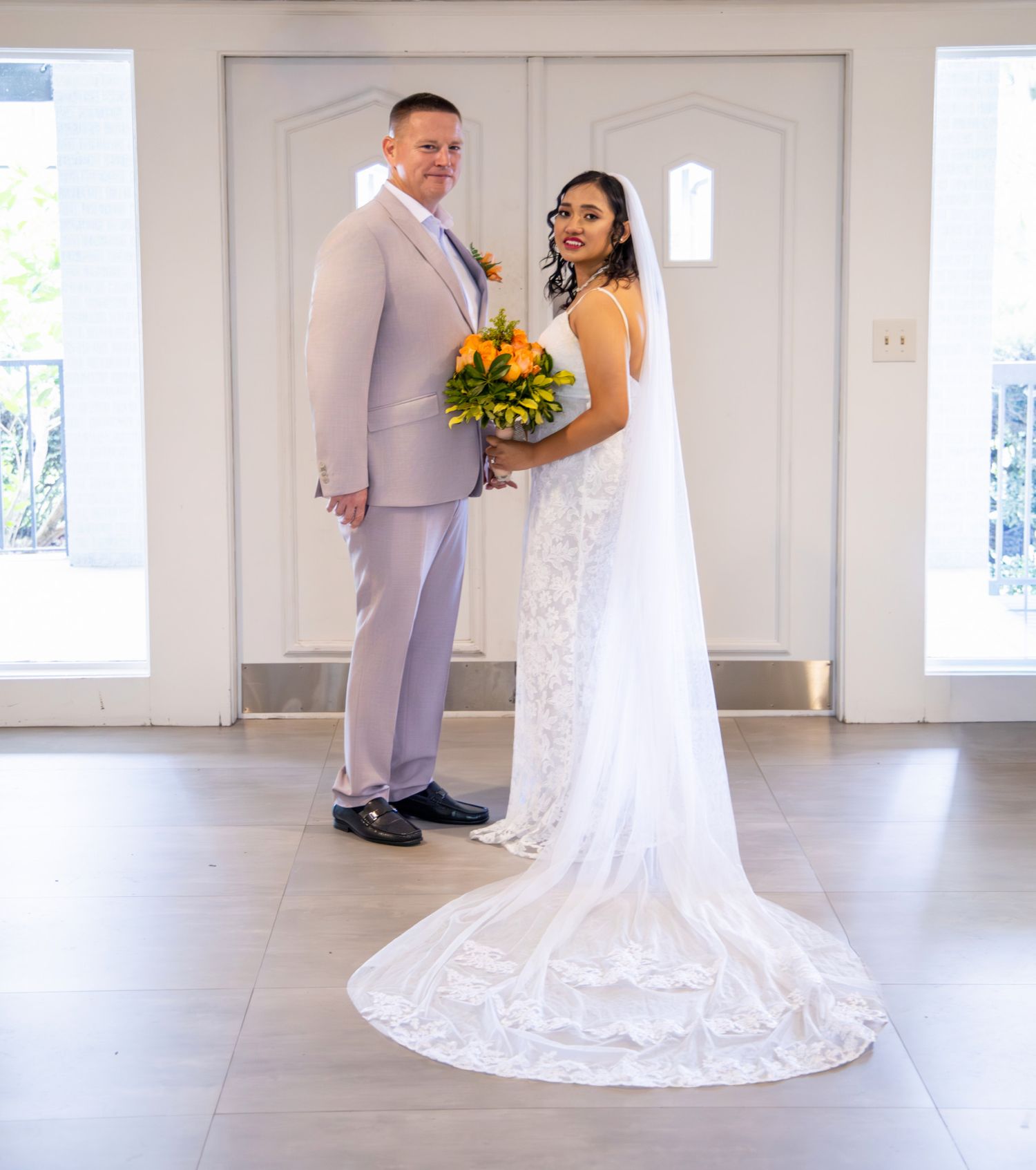 Bride in white lace gown holding orange bouquet stands with groom in lavender suit at elegant wedding venue.