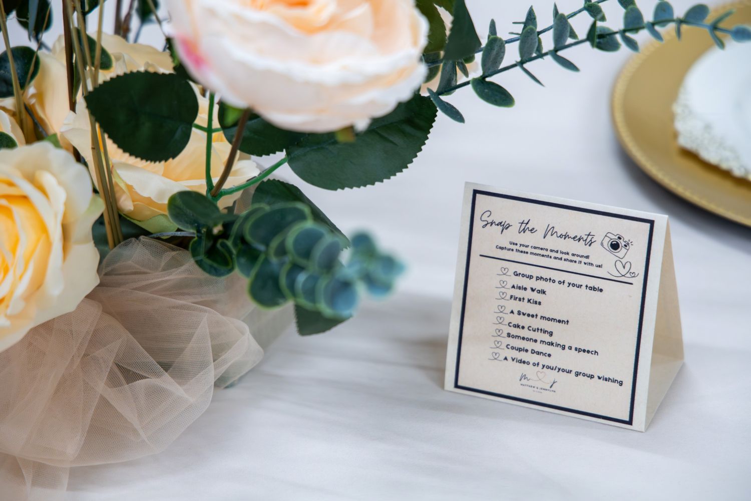 Elegant wedding table decor with white and pink roses, greenery, and a printed card on white linen.