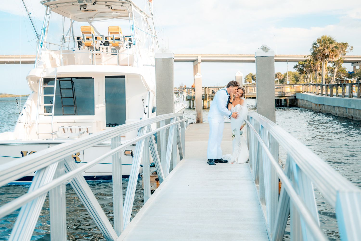 Couple embracing on a white dock beside a luxury yacht on calm blue waters at a marina.