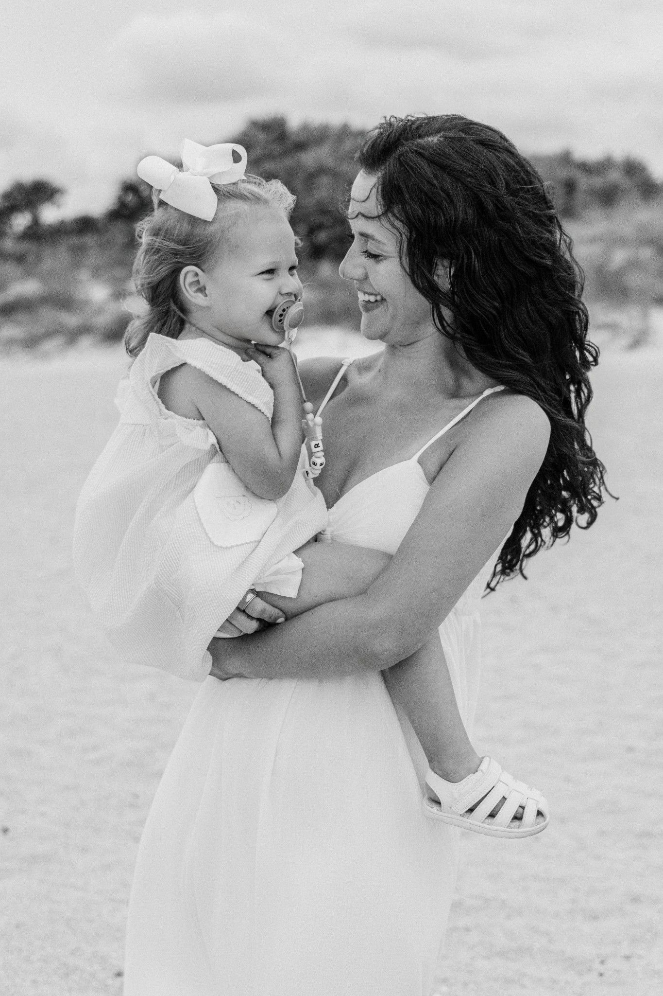 Black and white photo of a smiling mother in white dress holding her laughing little girl on a beach.