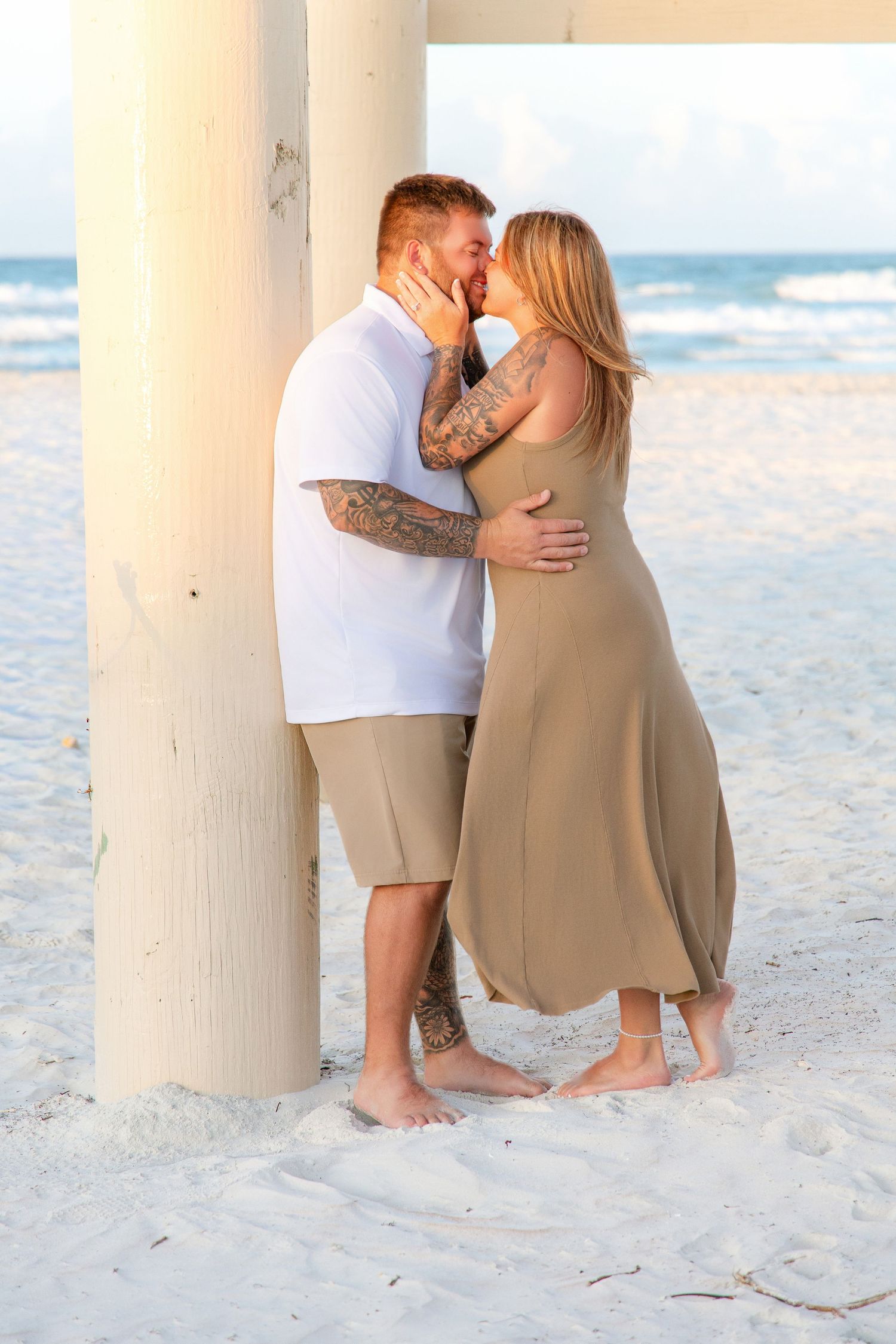 Couple with tattoos kissing barefoot on white sand beach near pier at golden hour.