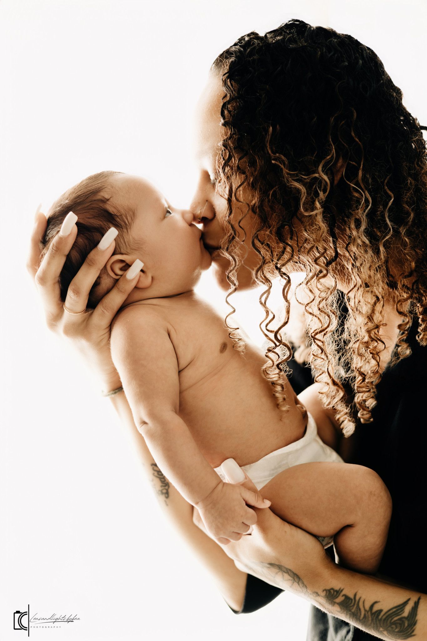 A tender moment captured in sepia tones shows curly-haired figure holding and kissing a newborn baby.