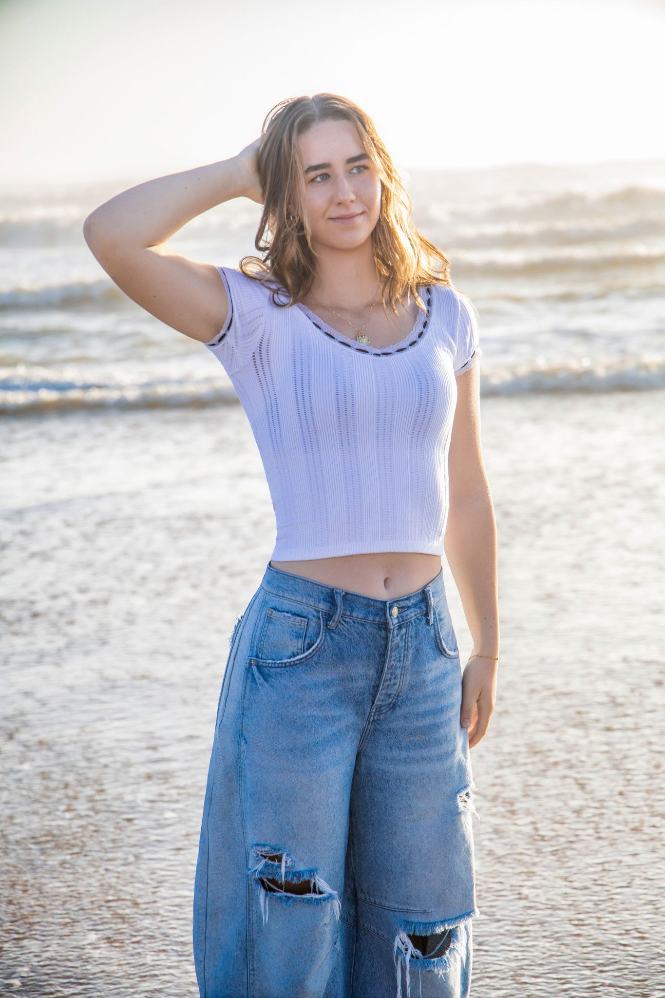 Person wearing a white crop top and ripped jeans poses on a beach during sunset.