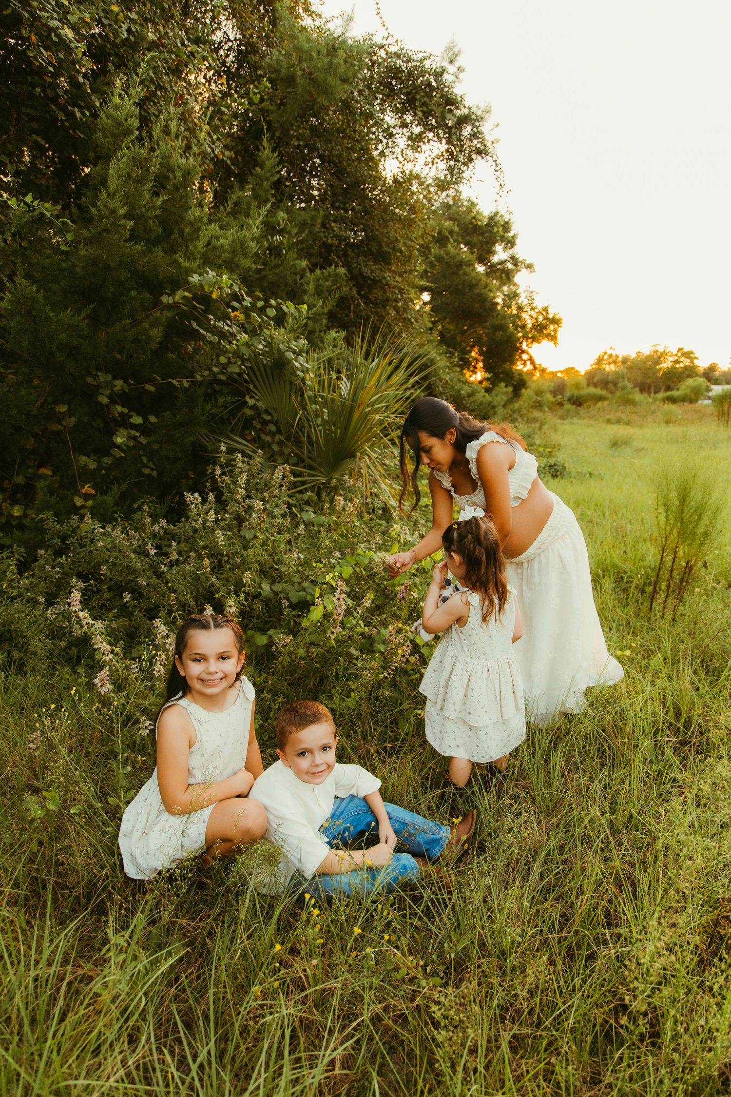 A mother and three children in white dresses play in a lush green field during golden hour sunset.