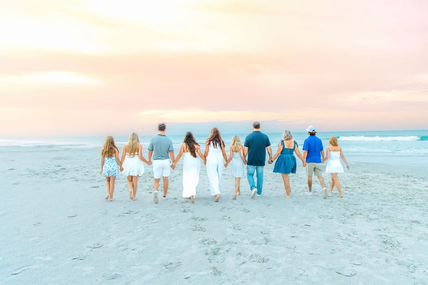 Large family group holding hands walking together on a sandy beach at sunset with pastel pink and blue sky.