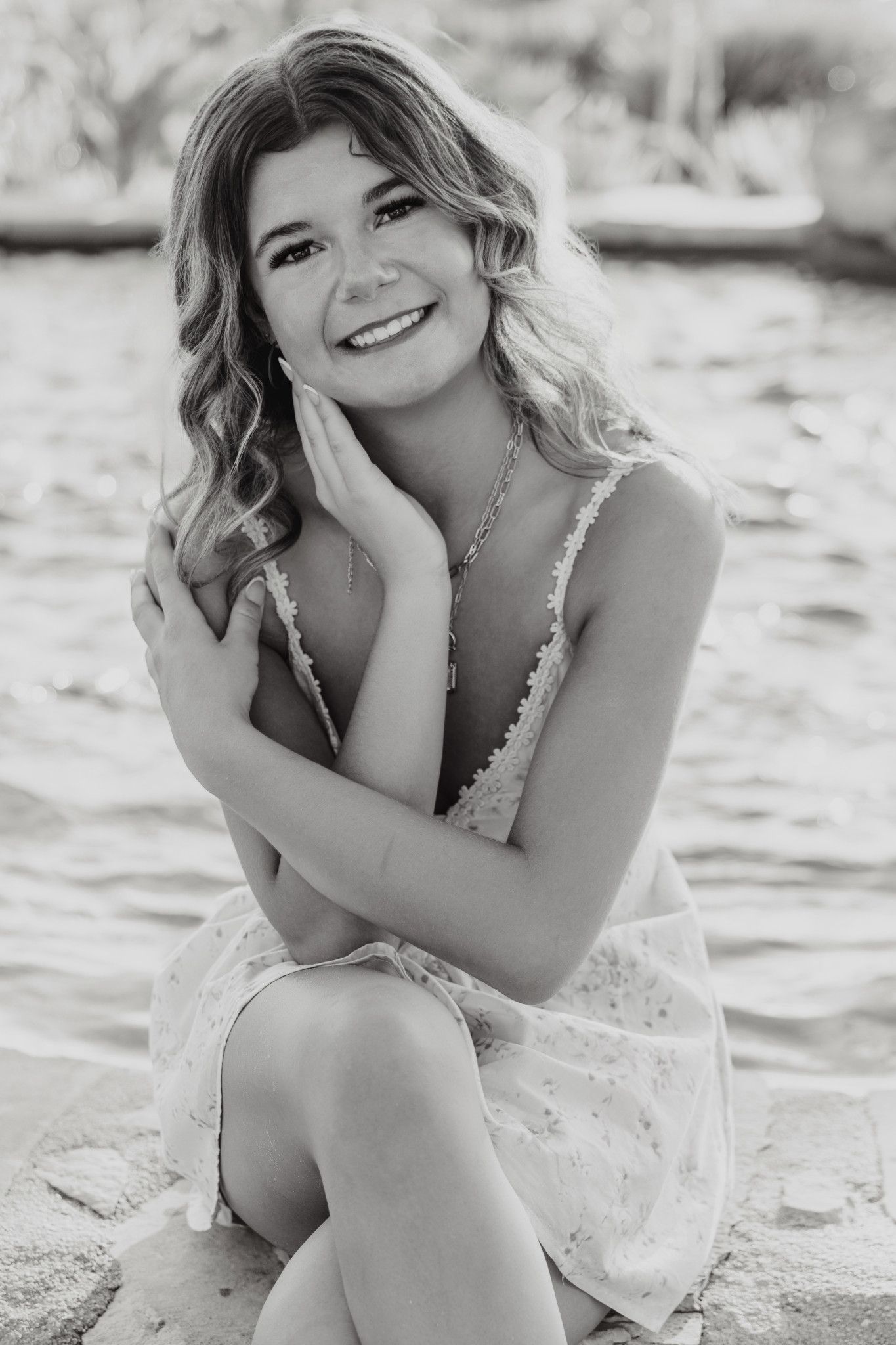 Black and white photo of a smiling young woman in a floral dress sitting on a sandy beach.