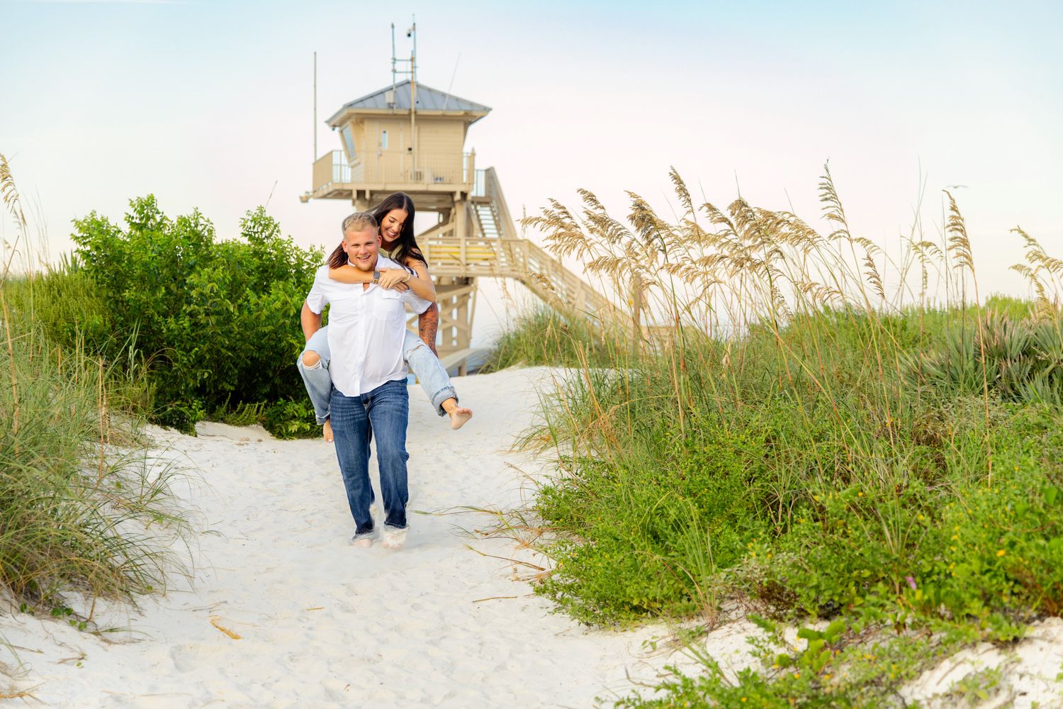 Couple enjoying a piggyback ride on white sand beach dunes with sea oats and a lifeguard tower backdrop.