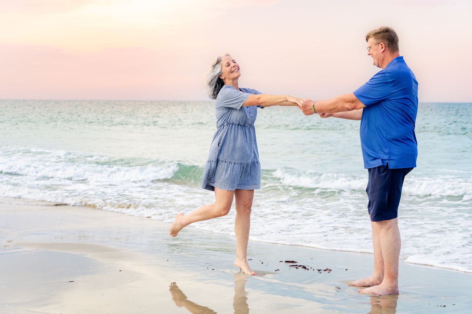 Happy couple dancing and holding hands on a sandy beach at sunset with ocean waves.