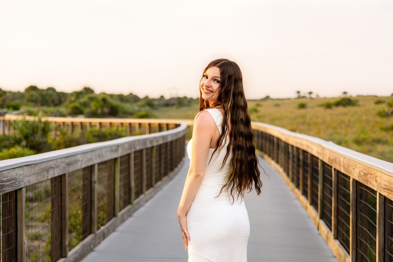 Young woman in white dress smiling on a wooden boardwalk at sunset.