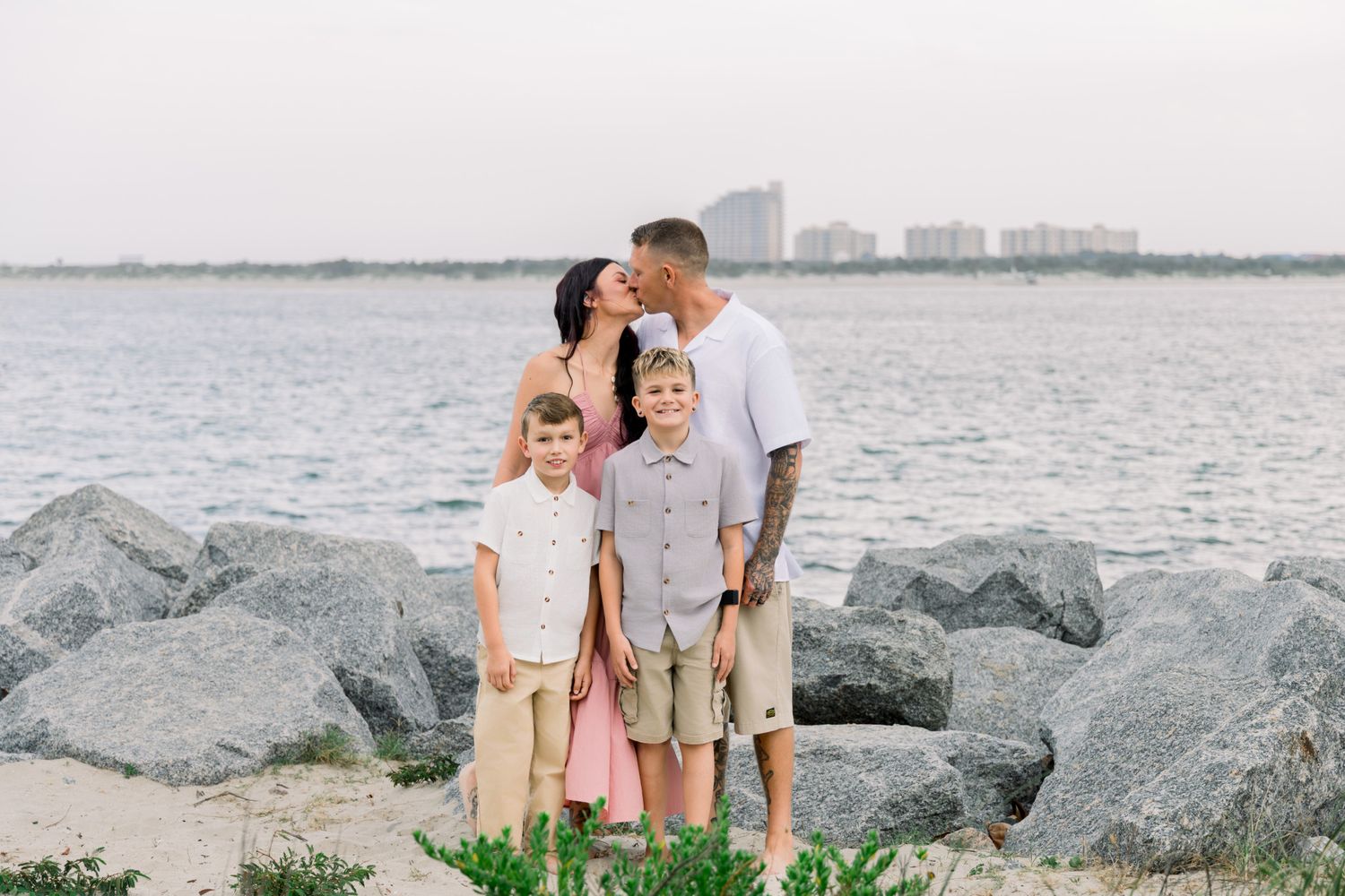 Family of four poses on rocky beach, parents kissing behind two young boys, city skyline and ocean in background.