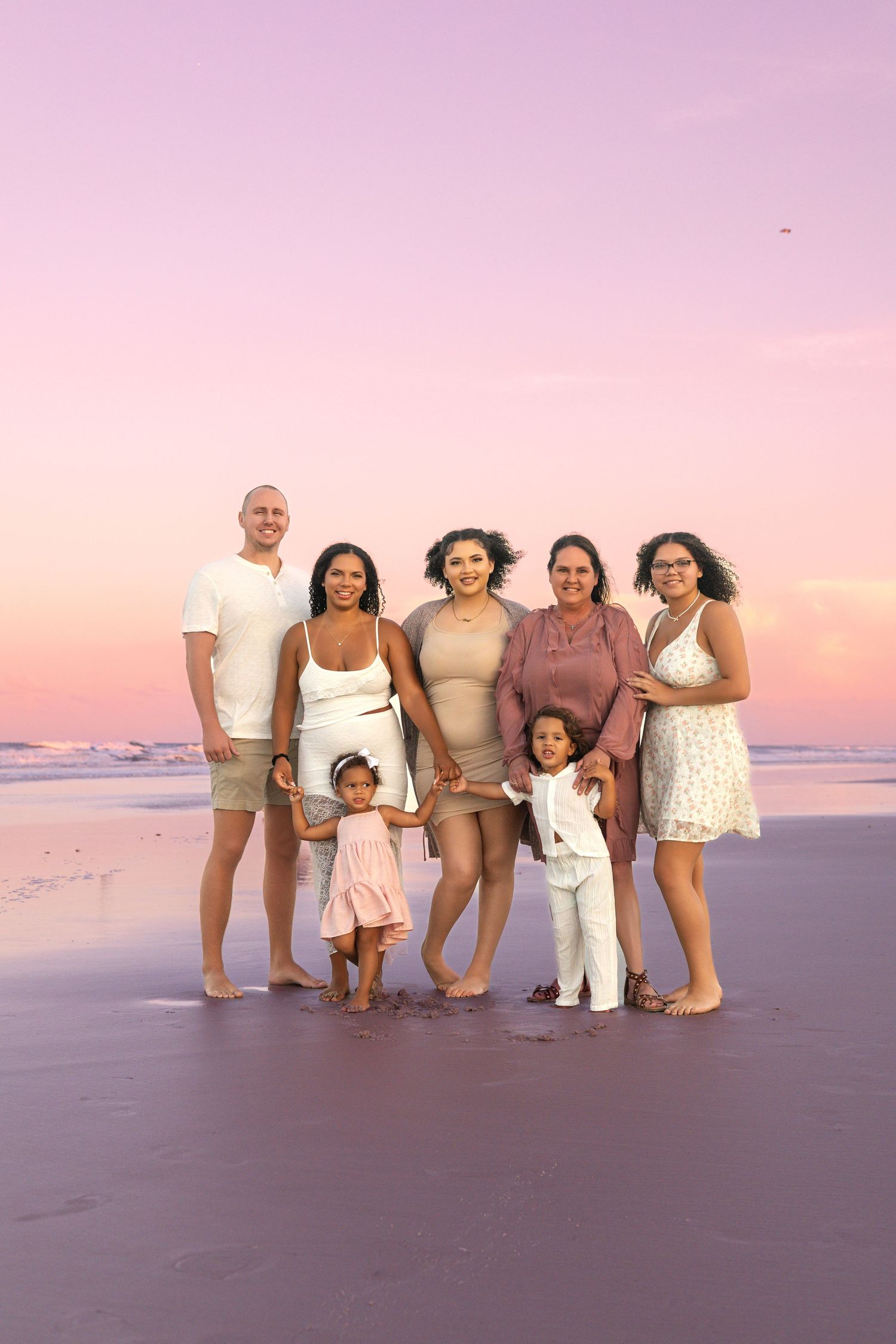 A family group poses together on the beach at sunset wearing coordinated white and neutral outfits.