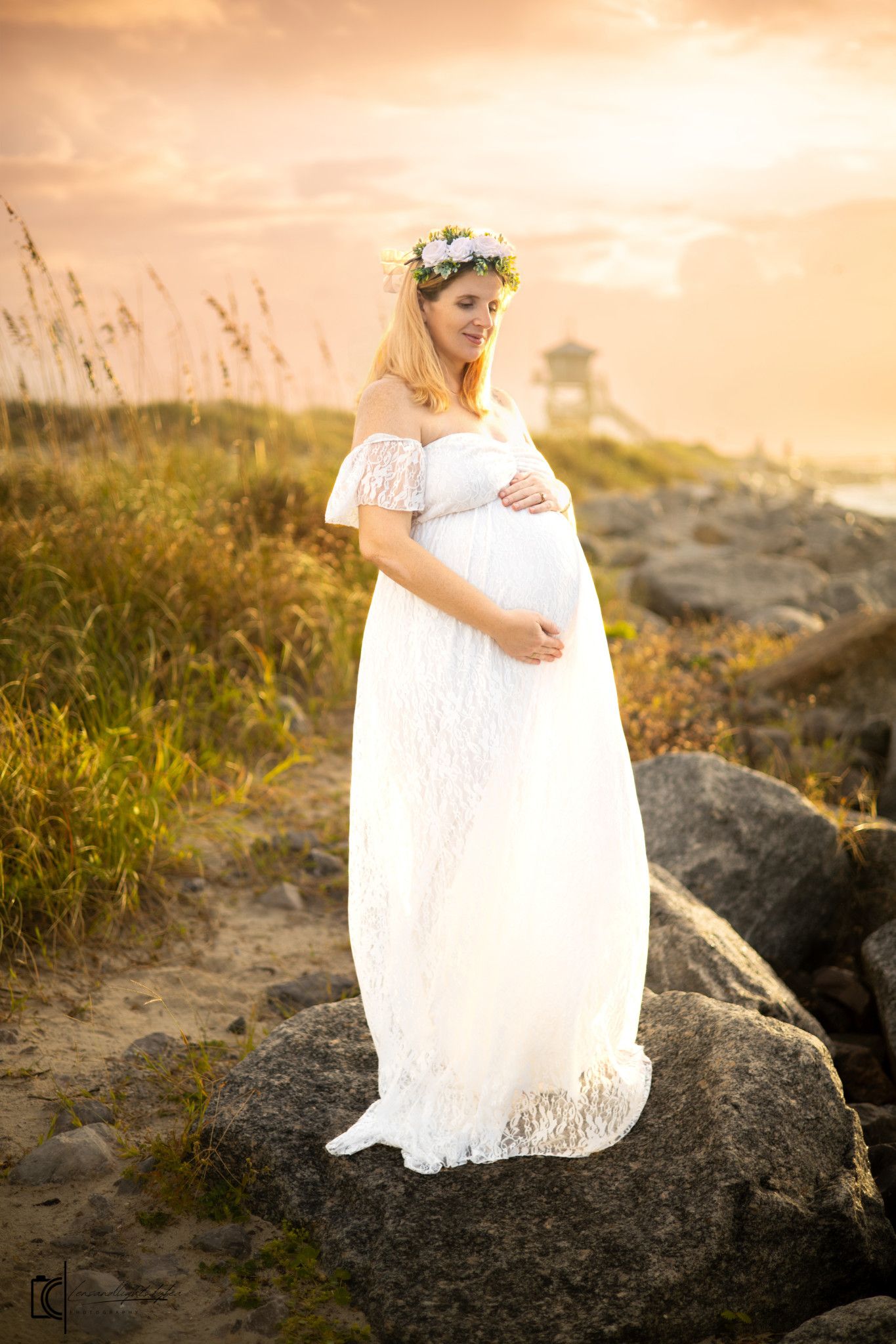 A figure in a flowing white lace dress sits on coastal rocks at sunset creating a dreamy silhouette against the golden sky.